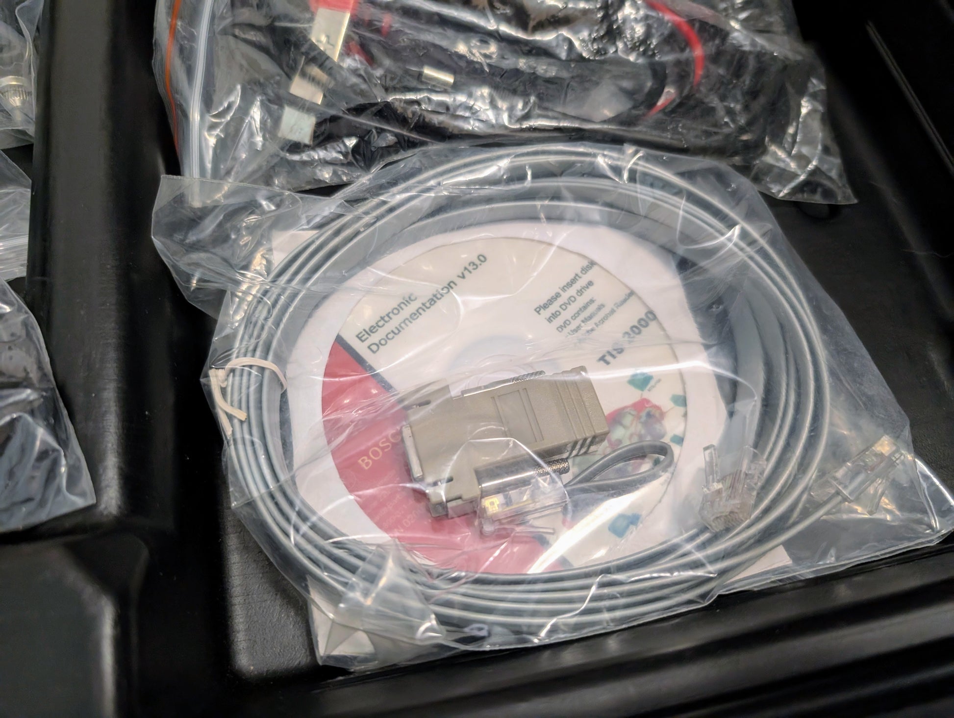 A sealed plastic bag holds a coiled gray serial cable, Electronic Documentation v3.0 CD, and extra connectors—all essentials for the Vetronix TESTED Tech 2 Automotive Diagnostic Tool for 1991-2013 GM vehicles—organized in a black tray.