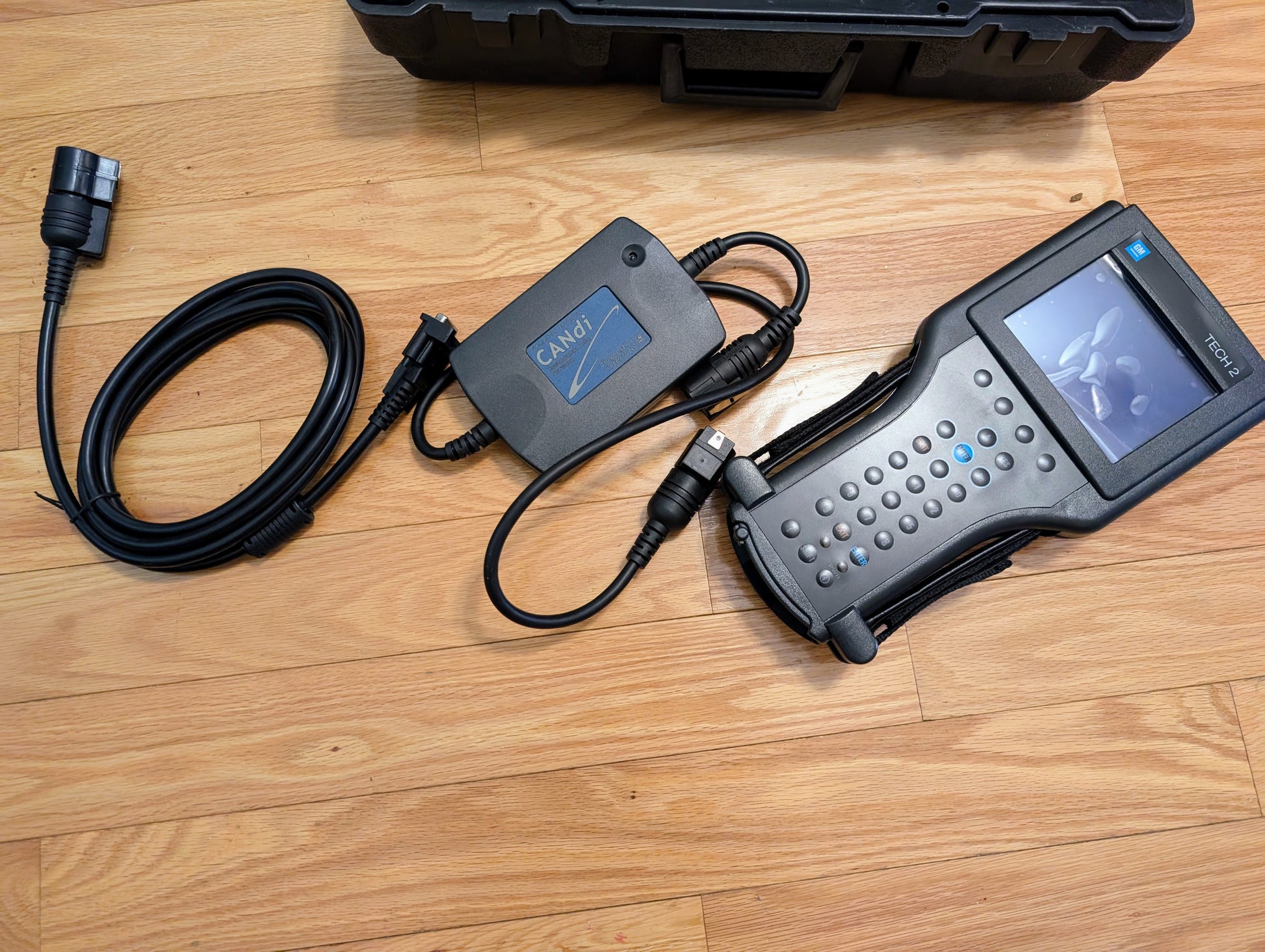 A Vetronix TESTED Tech 2 Automotive Diagnostic Tool & Accessories for 1991-2013 GM Vehicles, including a keypad, screen, cables, and CANdi module, is displayed on a wooden floor with a black carrying case partially visible above.
