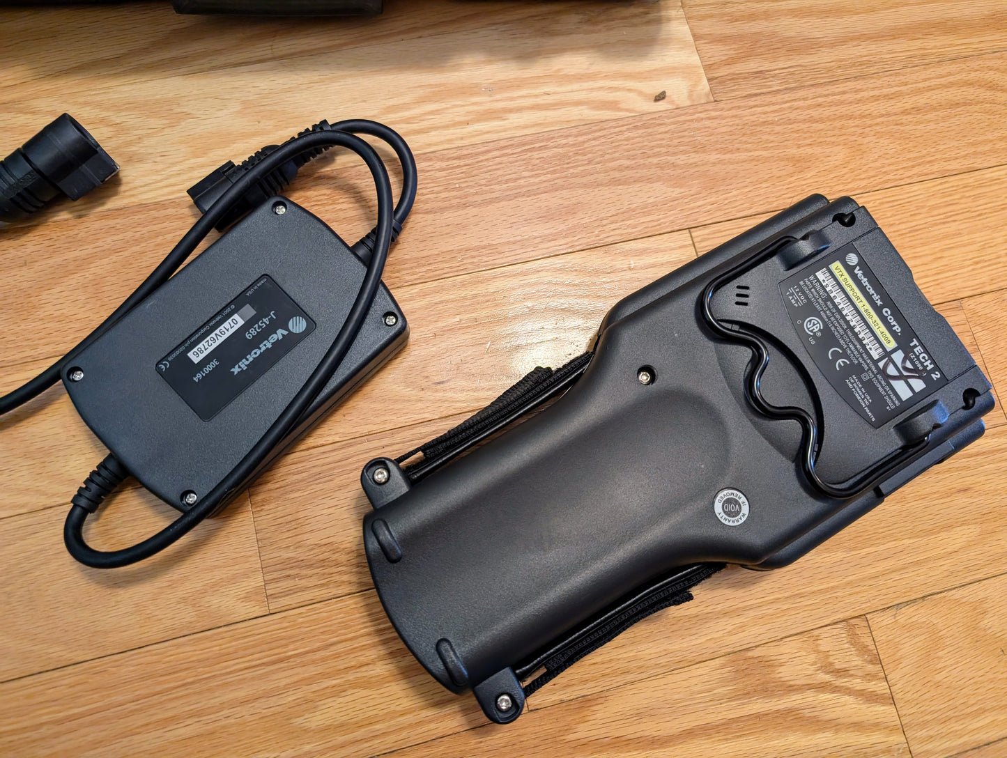 A Vetronix TESTED Tech 2 Automotive Diagnostic Tool for 1991-2013 GM vehicles with a holster is on a wooden floor beside its rectangular black charger with attached cables.