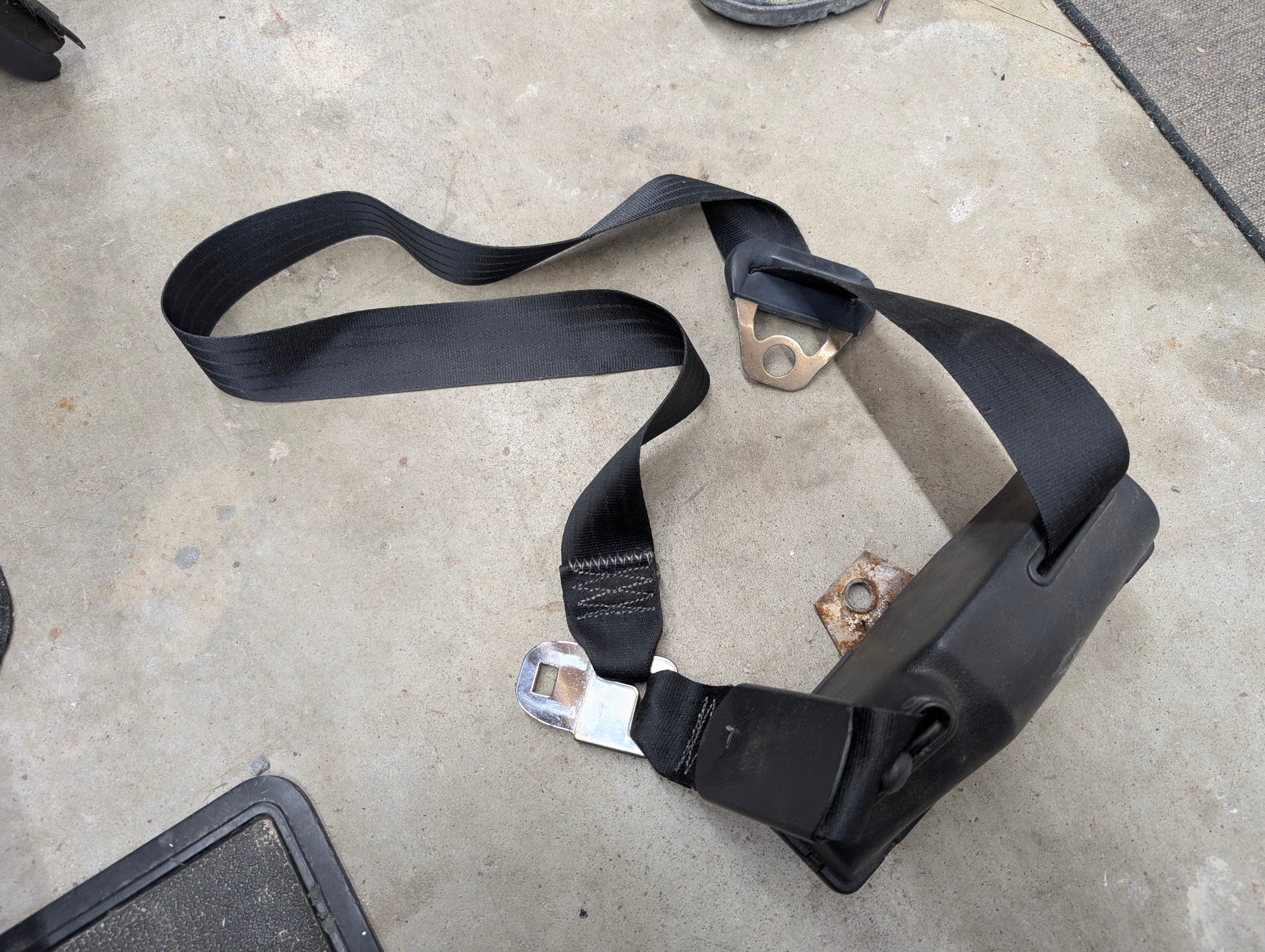 A General Motors NOS Passenger Seat Belt Retractor in Charcoal for 1982-1993 Chevy S10, Blazer, and more, with metal buckle and brackets, is partially extended and twisted near its casing on a concrete floor.