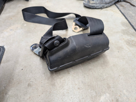 A NOS General Motors passenger seat belt retractor in charcoal for 1982-1993 Chevy S10, Blazer, and more rests on concrete. A boot is partially visible in the background.