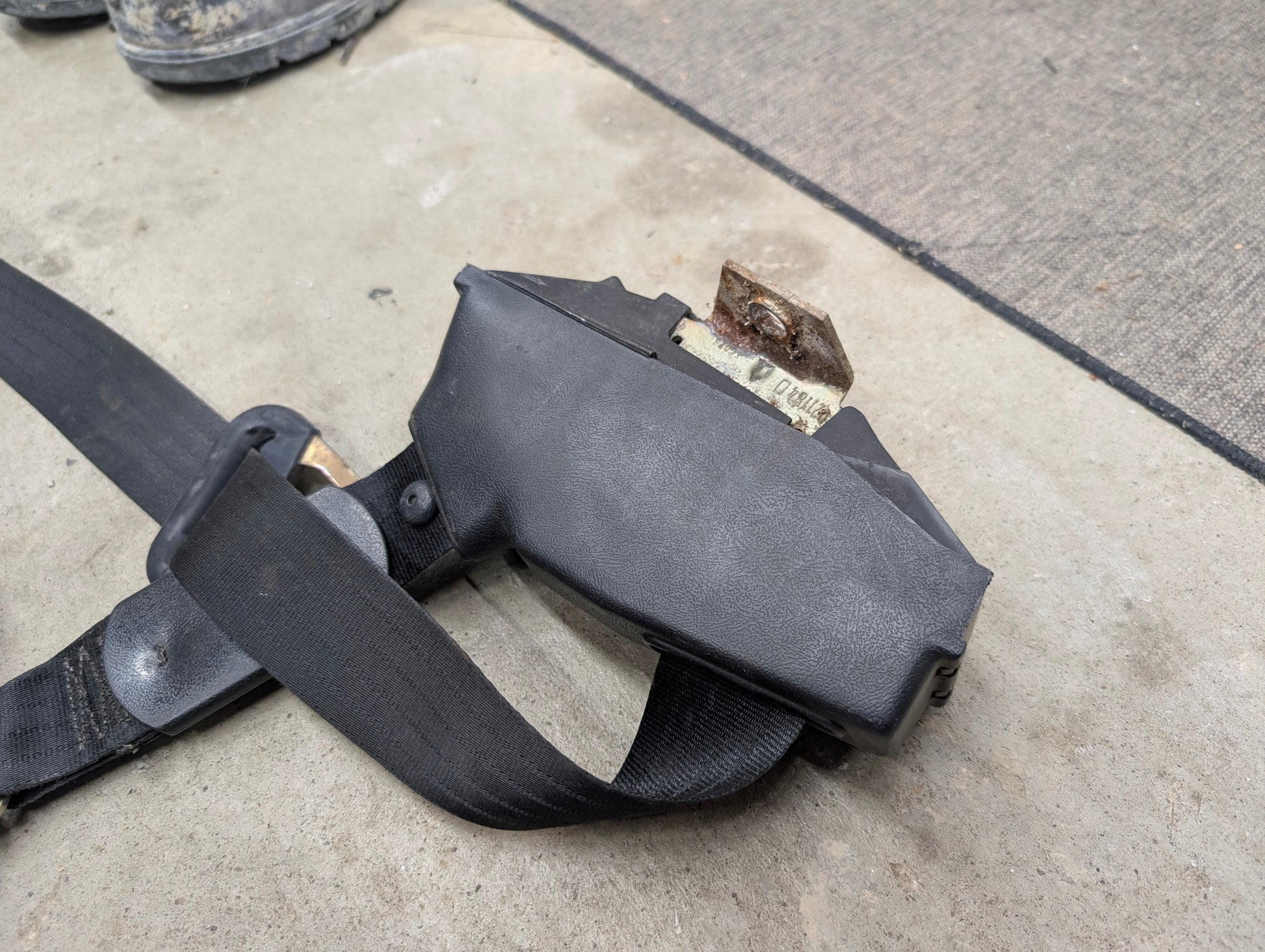 A NOS General Motors passenger seat belt retractor in charcoal for 1982-1993 Chevy S10, Blazer, and more, shown on a concrete surface.