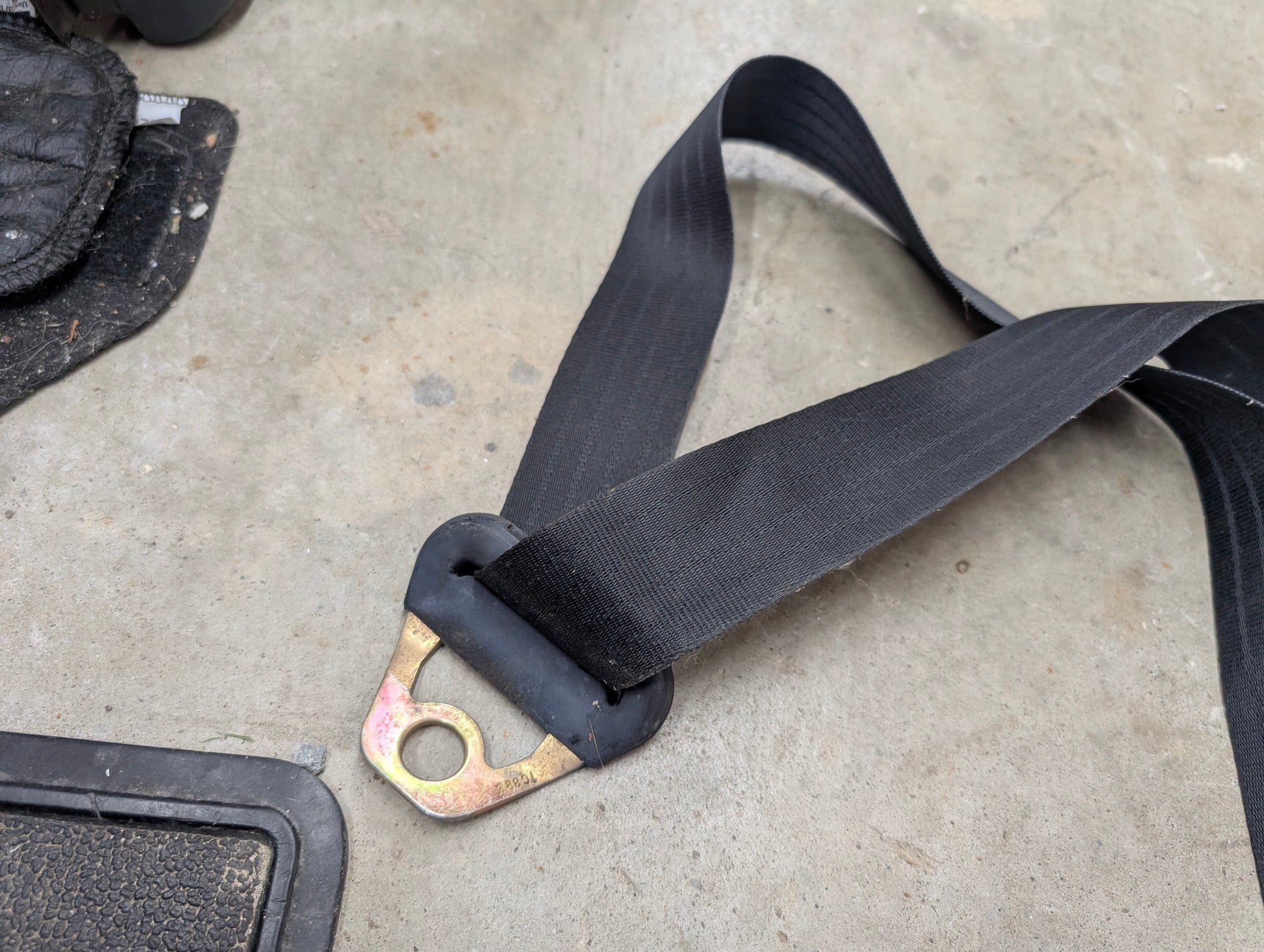 A General Motors NOS Passenger Seat Belt Retractor in charcoal for 1982-1993 Chevy S10 and Blazer rests on a gray concrete floor by the corner of a matching Chevrolet S10 floor mat.