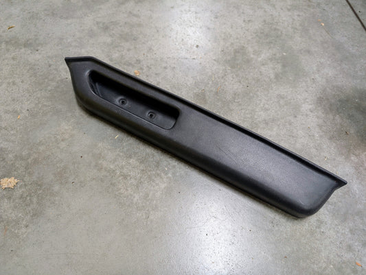 A General Motors OEM black passenger door panel armrest handle for 1986-1993 Chevy S10 Blazer, GMC Jimmy, and more rests on a gray concrete floor. The handle is rectangular with angular edges, recessed grip area, and two screw holes.