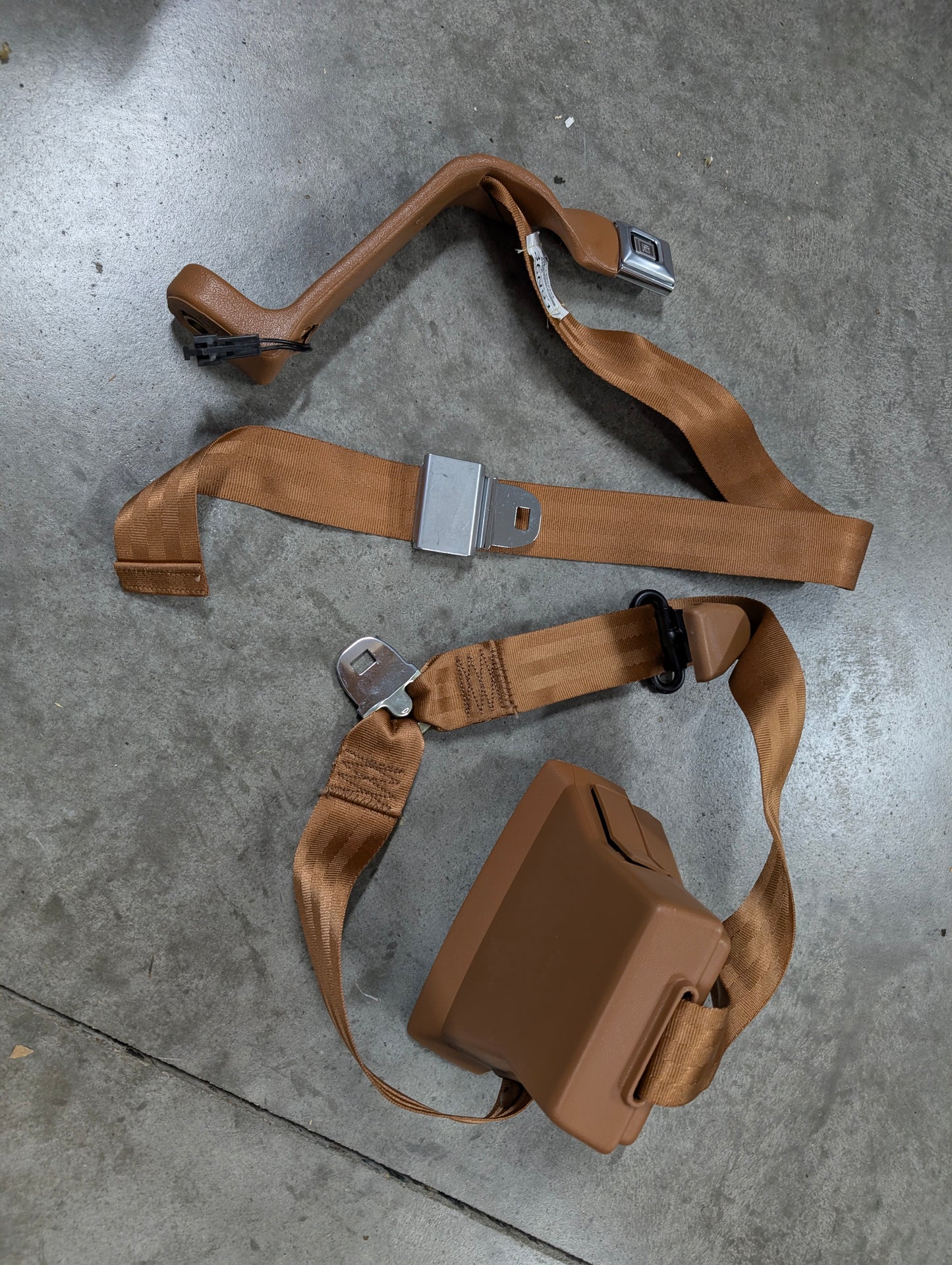 A General Motors NOS passenger seat belt retractor and buckle in brown for 1982-1993 Chevy S10 and GMC S15 is displayed on a gray concrete floor, highlighting its classic automotive design.