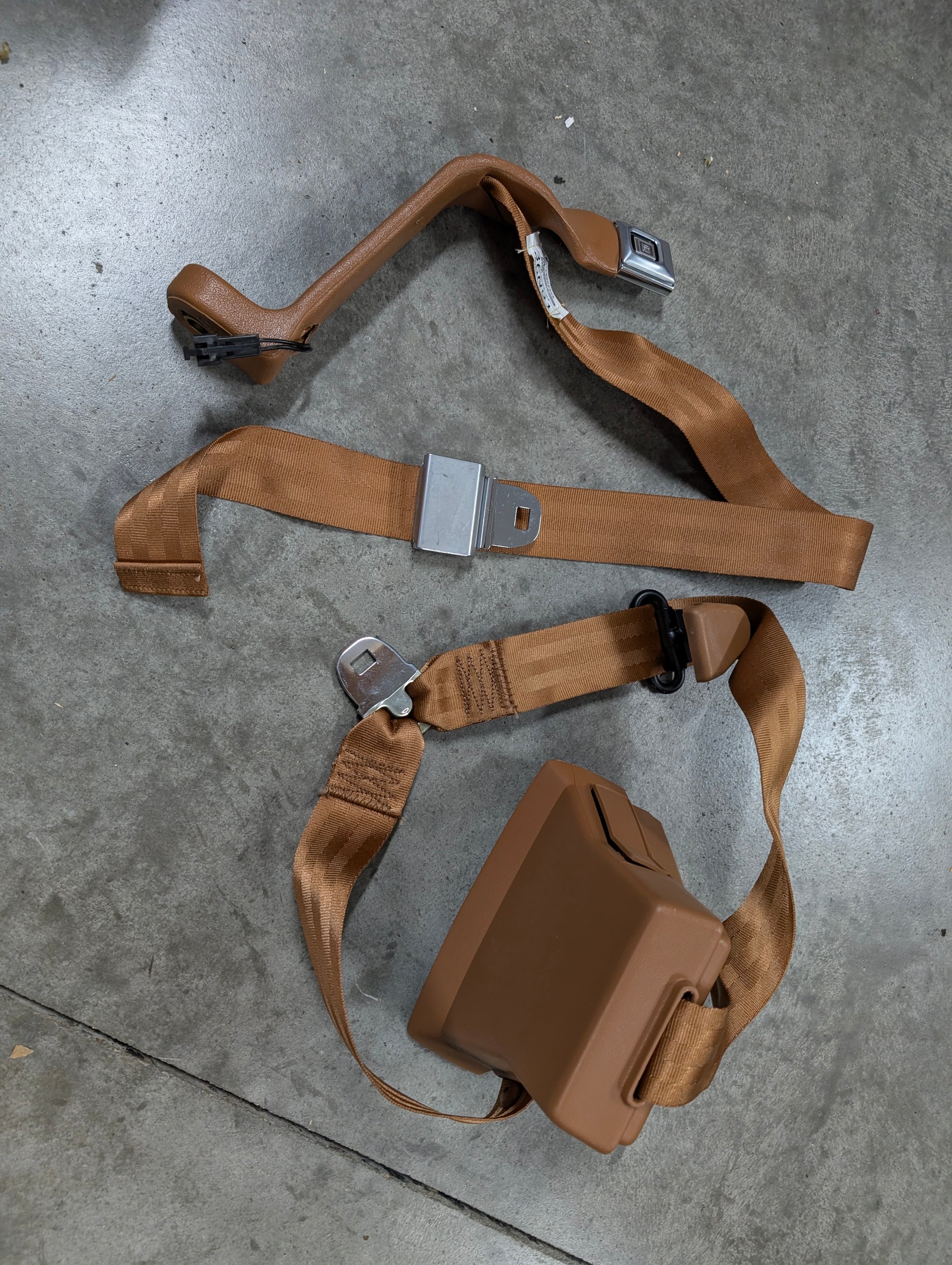 A General Motors NOS passenger seat belt retractor and buckle in brown for 1982-1993 Chevy S10 and GMC S15 is displayed on a gray concrete floor, highlighting its classic automotive design.