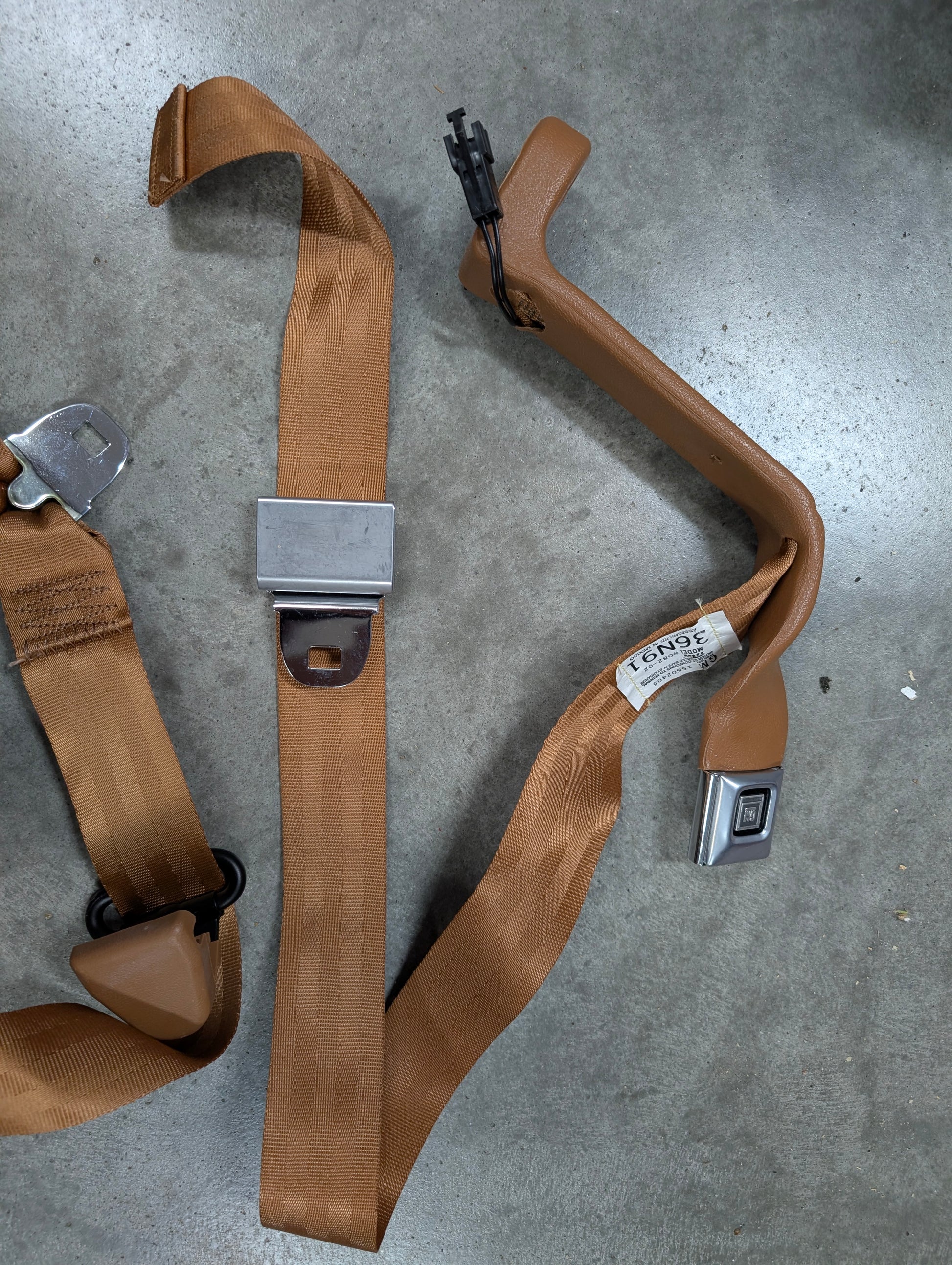 NOS General Motors brown passenger seat belt retractor & buckle for 1982-1993 Chevy S10/GMC S15, laid out on a gray concrete surface.
