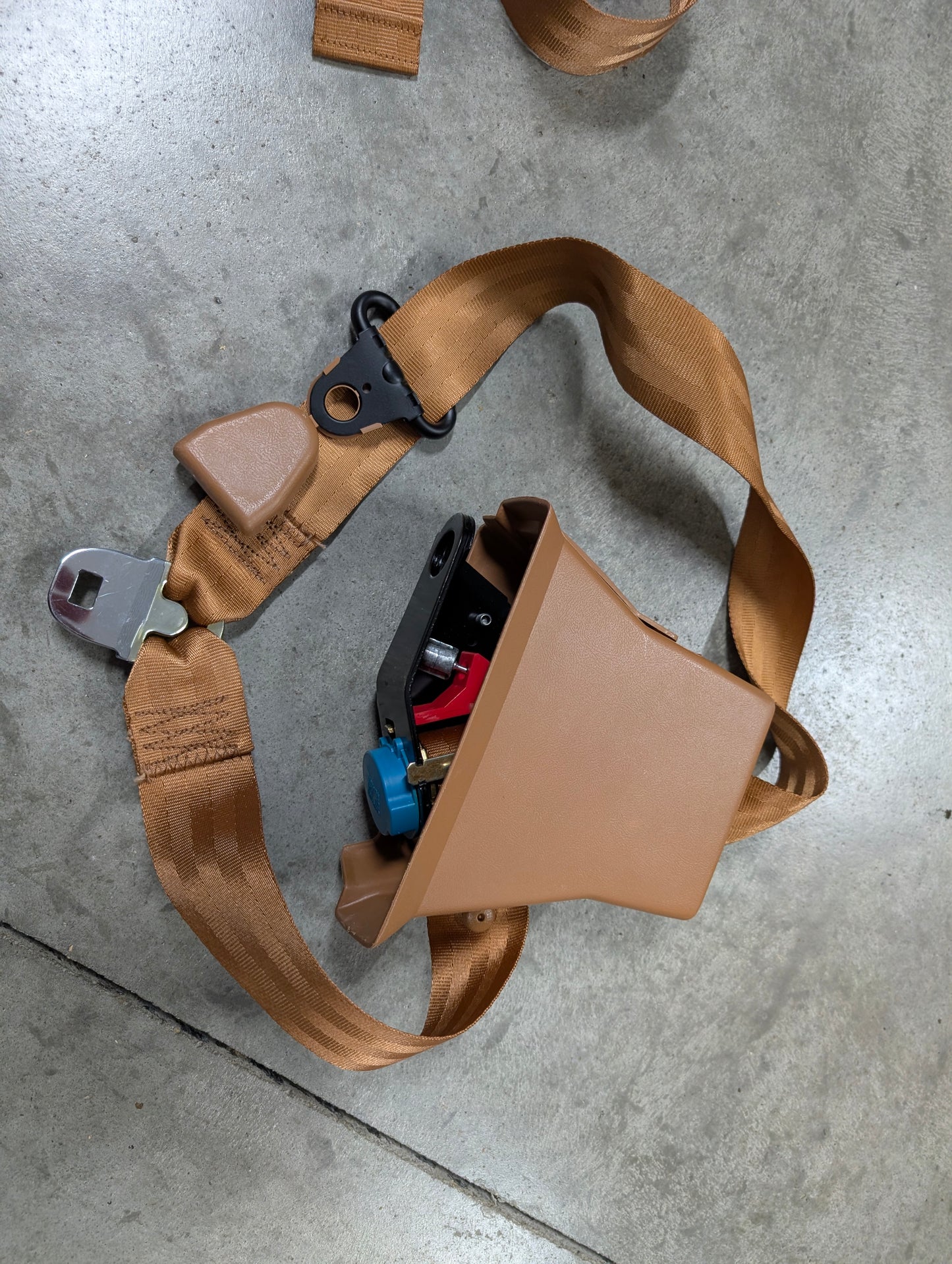 A General Motors NOS Passenger Seat Belt Retractor & Buckle in brown for 1982-1993 Chevy S10, GMC S15 rests on a gray concrete floor, featuring a plastic pouch holding tools and keys.