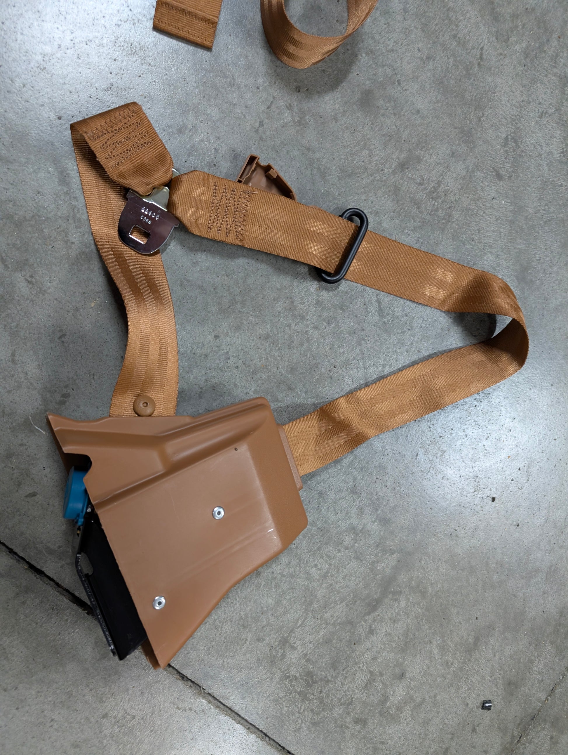 A General Motors NOS Passenger Seat Belt Retractor & Buckle in brown for 1982-1993 Chevy S10, GMC S15 is shown on a concrete floor, featuring tan webbing, black metal hardware, and a quick-release buckle.