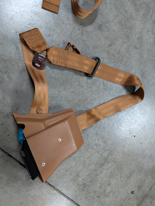 A General Motors NOS Passenger Seat Belt Retractor & Buckle in brown for 1982-1993 Chevy S10, GMC S15 is shown on a concrete floor, featuring tan webbing, black metal hardware, and a quick-release buckle.