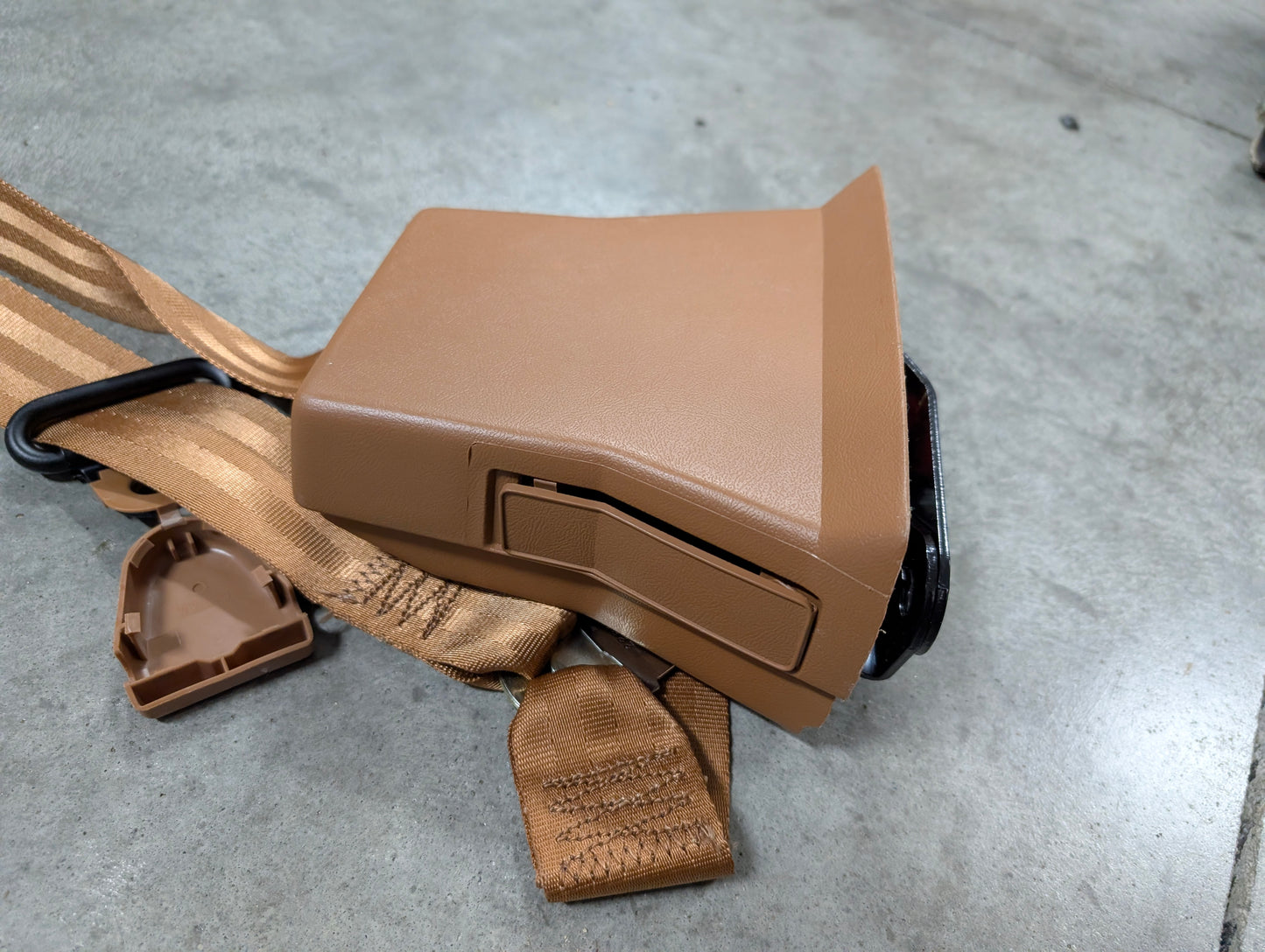 A General Motors NOS brown passenger seat belt retractor & buckle for 1982–1993 Chevy S10/GMC S15 lies on a gray concrete floor, with the plastic cover partially detached to reveal part of the mechanism.