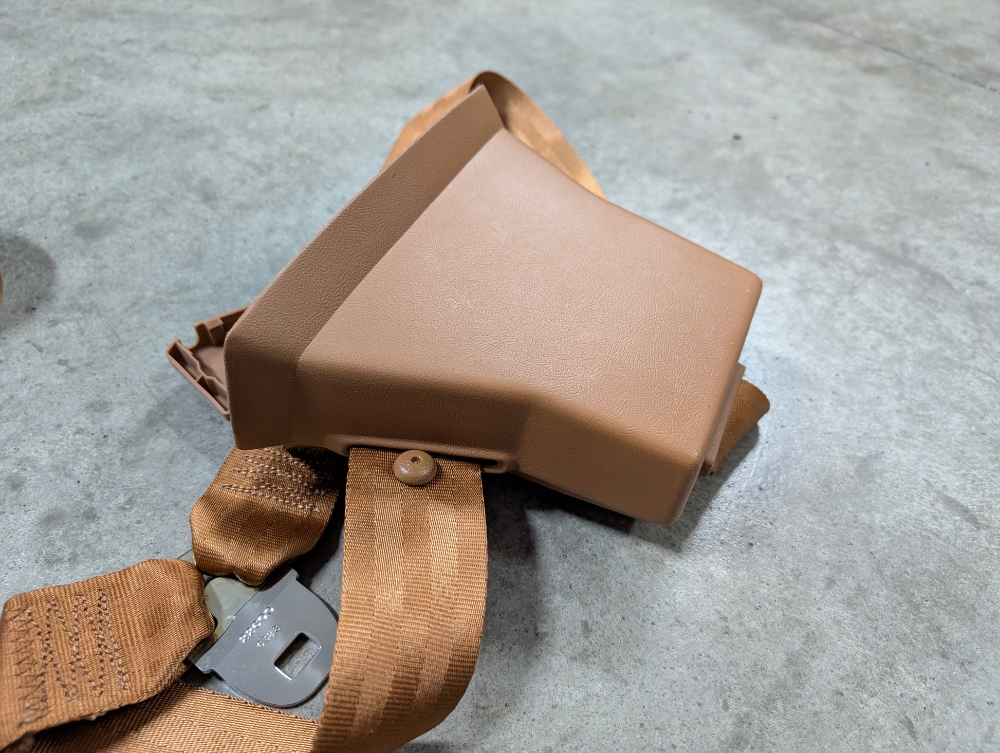 A General Motors NOS brown passenger seat belt retractor and buckle for 1982-1993 Chevy S10/GMC S15 lies on a gray concrete floor, with the metal latch visible beside it.