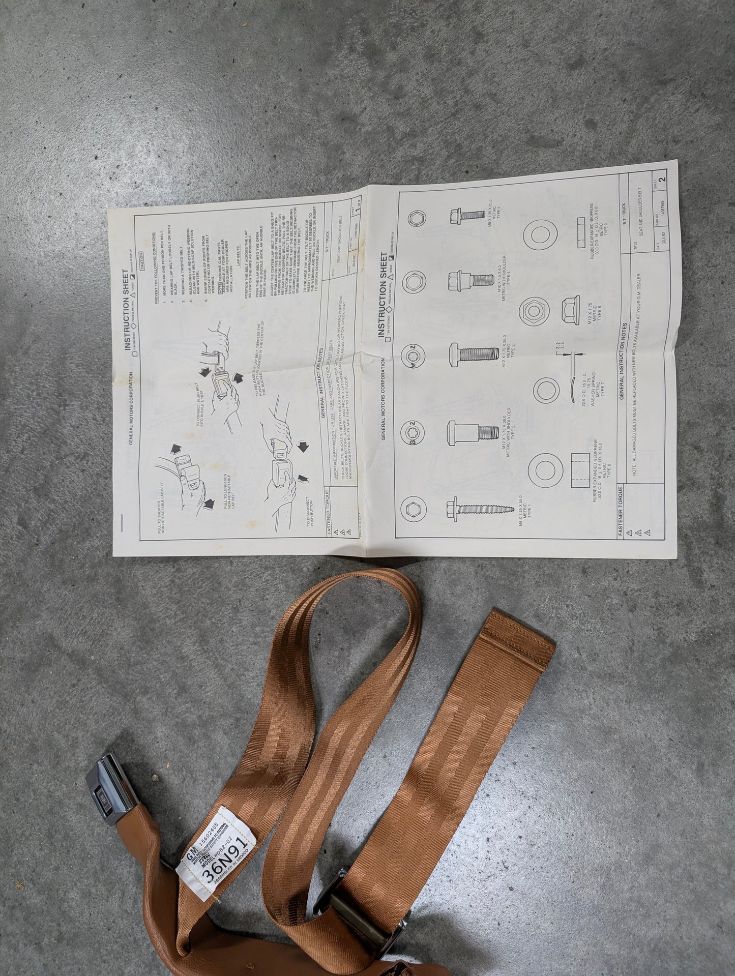 A General Motors NOS passenger seat belt retractor & buckle in brown for 1982–1993 Chevy S10/GMC S15 and technical instructions with bolt diagrams are spread out on a gray concrete floor.