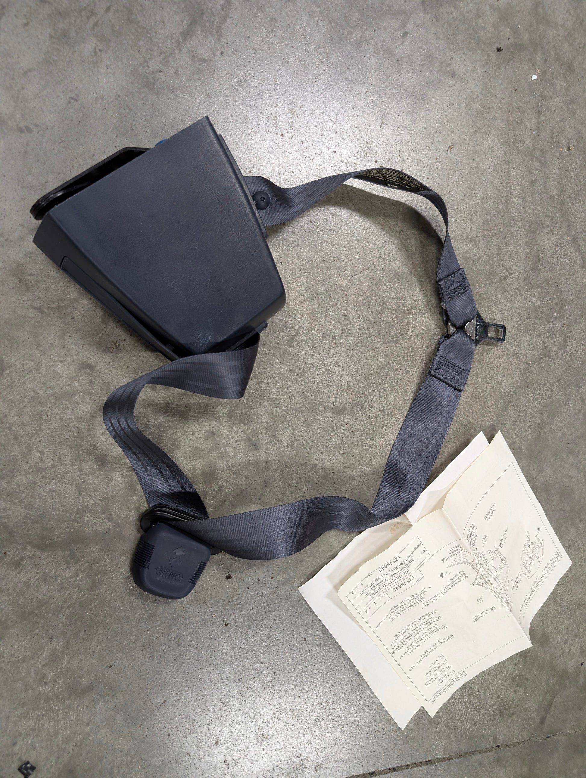 A NOS blue passenger seat belt retractor and buckle by General Motors for 1988–1994 Chevy/GMC C/K Trucks is displayed on a concrete floor beside printed instructions.