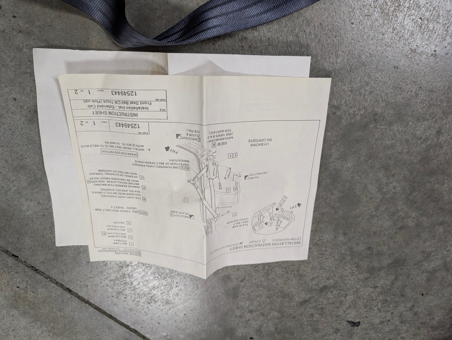 A sheet of assembly instructions with diagrams and text lies on a gray concrete floor. At the top left corner, a blue General Motors NOS Passenger Seat Belt Retractor & Buckle for 1988-1994 Chevy/GMC C/K Trucks is partially visible.