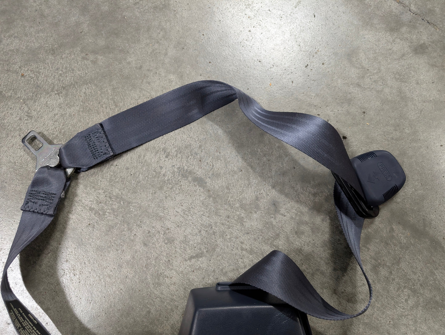 A close-up of a General Motors NOS blue passenger seat belt retractor and buckle for 1988-1994 Chevy/GMC C/K Trucks, lying on a smooth gray concrete floor.