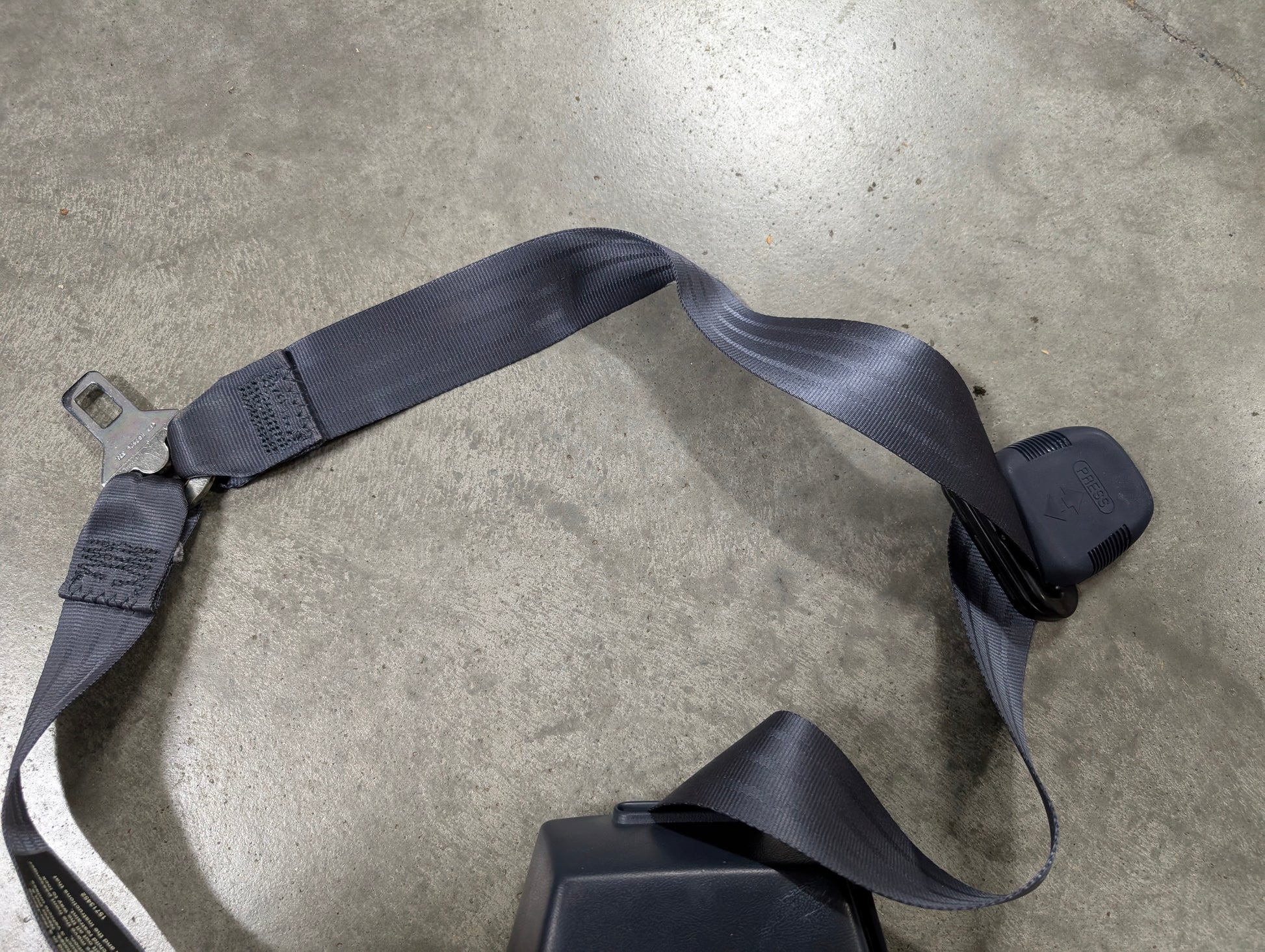 A close-up of a General Motors NOS blue passenger seat belt retractor and buckle for 1988-1994 Chevy/GMC C/K Trucks, lying on a smooth gray concrete floor.