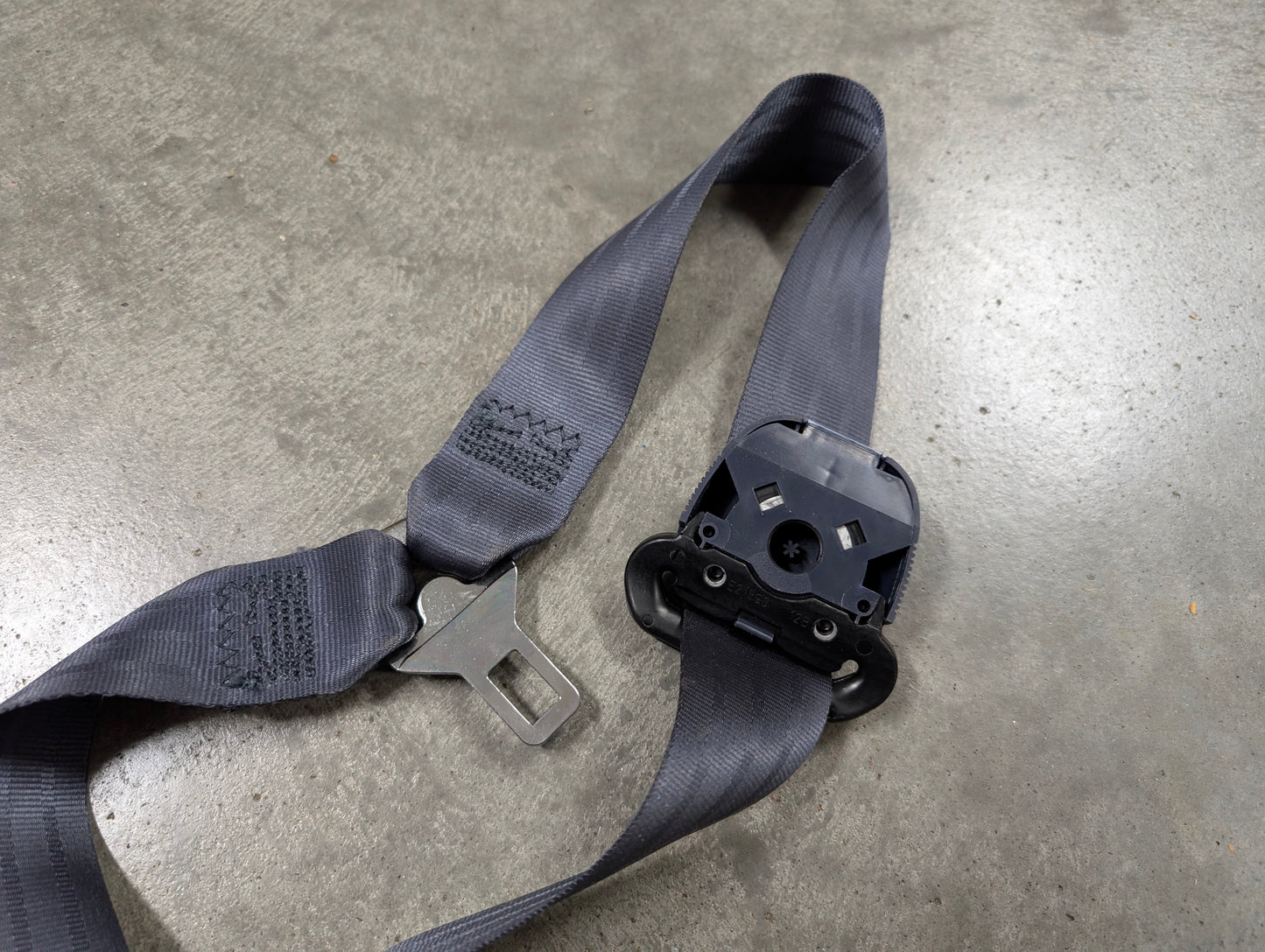 A blue General Motors NOS passenger seat belt retractor and buckle for 1988-1994 Chevy/GMC C/K trucks lies on a smooth concrete floor.