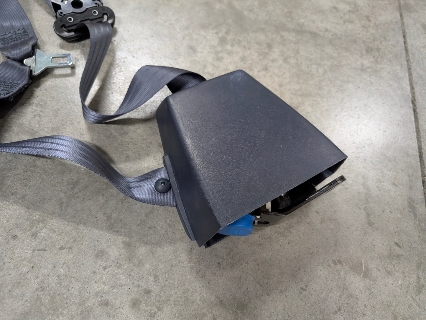 A close-up of a General Motors NOS Passenger Seat Belt Retractor & Buckle in Blue for 1988-1994 Chevy/GMC C/K Trucks, partially exposed and lying on a gray concrete floor.