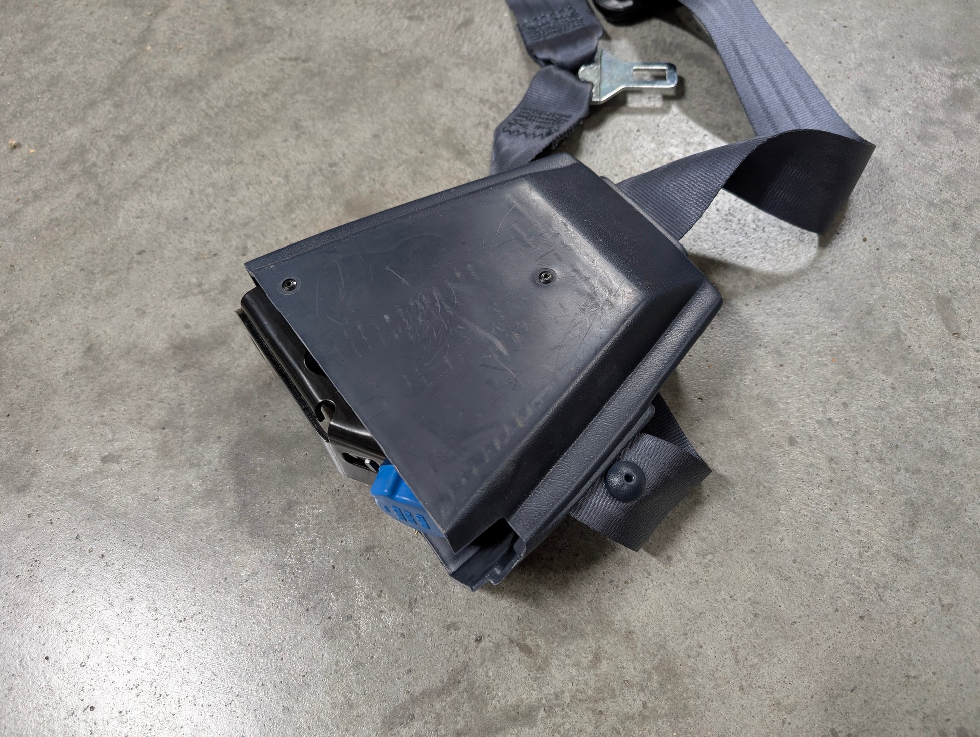 A NOS Passenger Seat Belt Retractor & Buckle in Blue for 1988-1994 Chevy/GMC C/K Trucks by General Motors is shown close-up on a concrete surface.