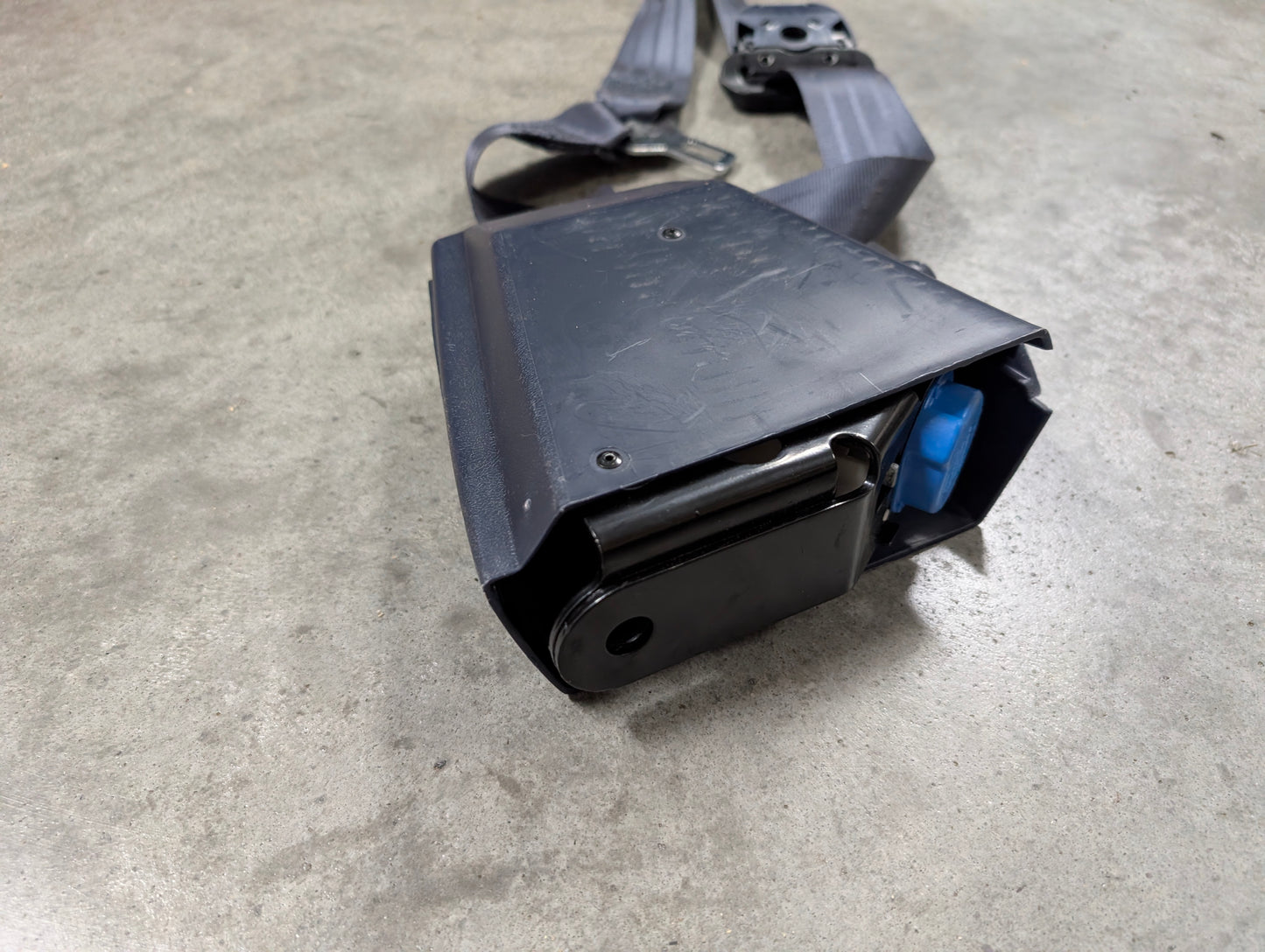 A close-up of a General Motors NOS Passenger Seat Belt Retractor & Buckle in Blue for 1988–1994 Chevy/GMC C/K Trucks, shown on a gray concrete floor with visible metal spool and blue OEM seat belt component.