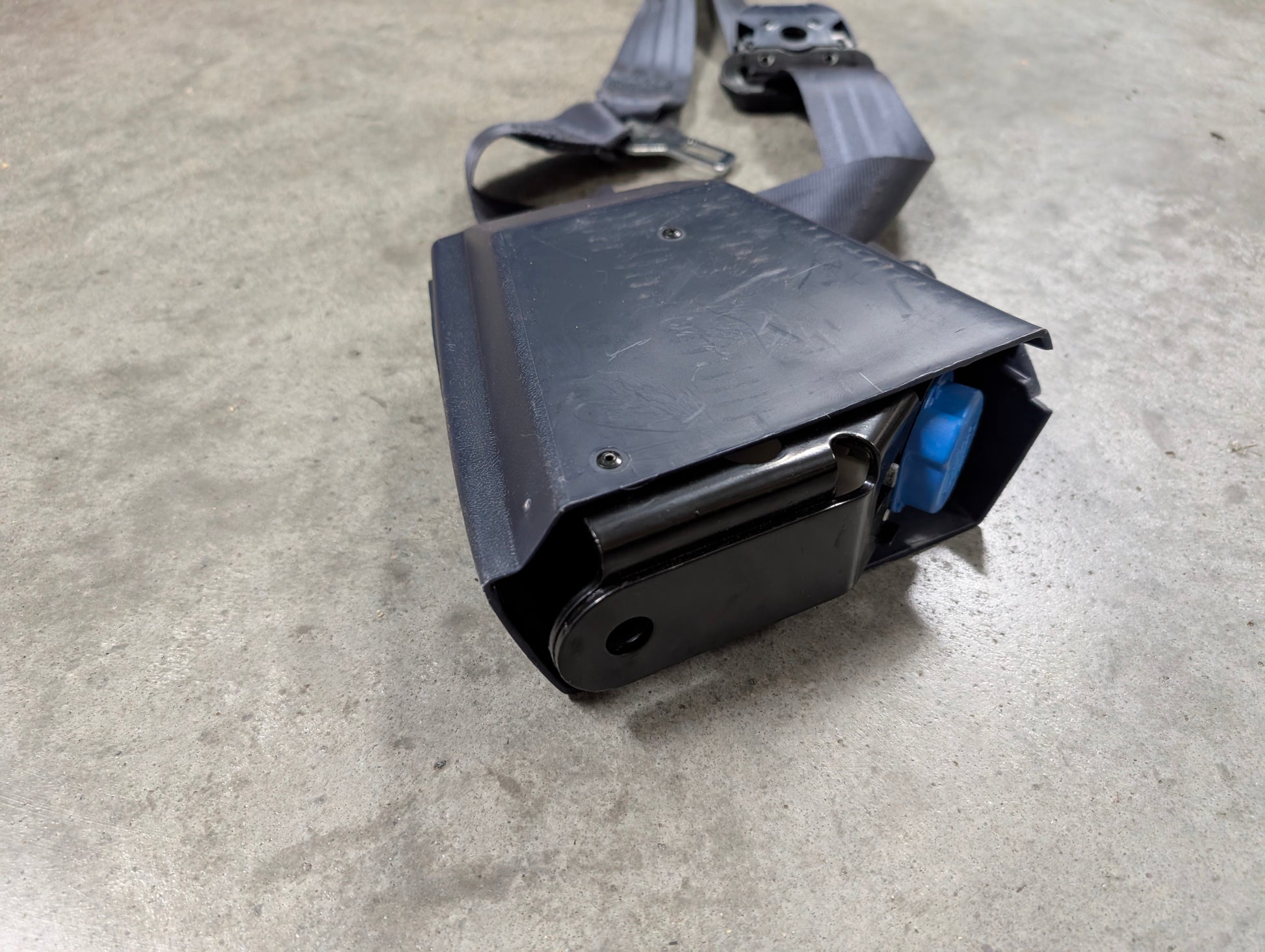 A close-up of a General Motors NOS Passenger Seat Belt Retractor & Buckle in Blue for 1988–1994 Chevy/GMC C/K Trucks, shown on a gray concrete floor with visible metal spool and blue OEM seat belt component.