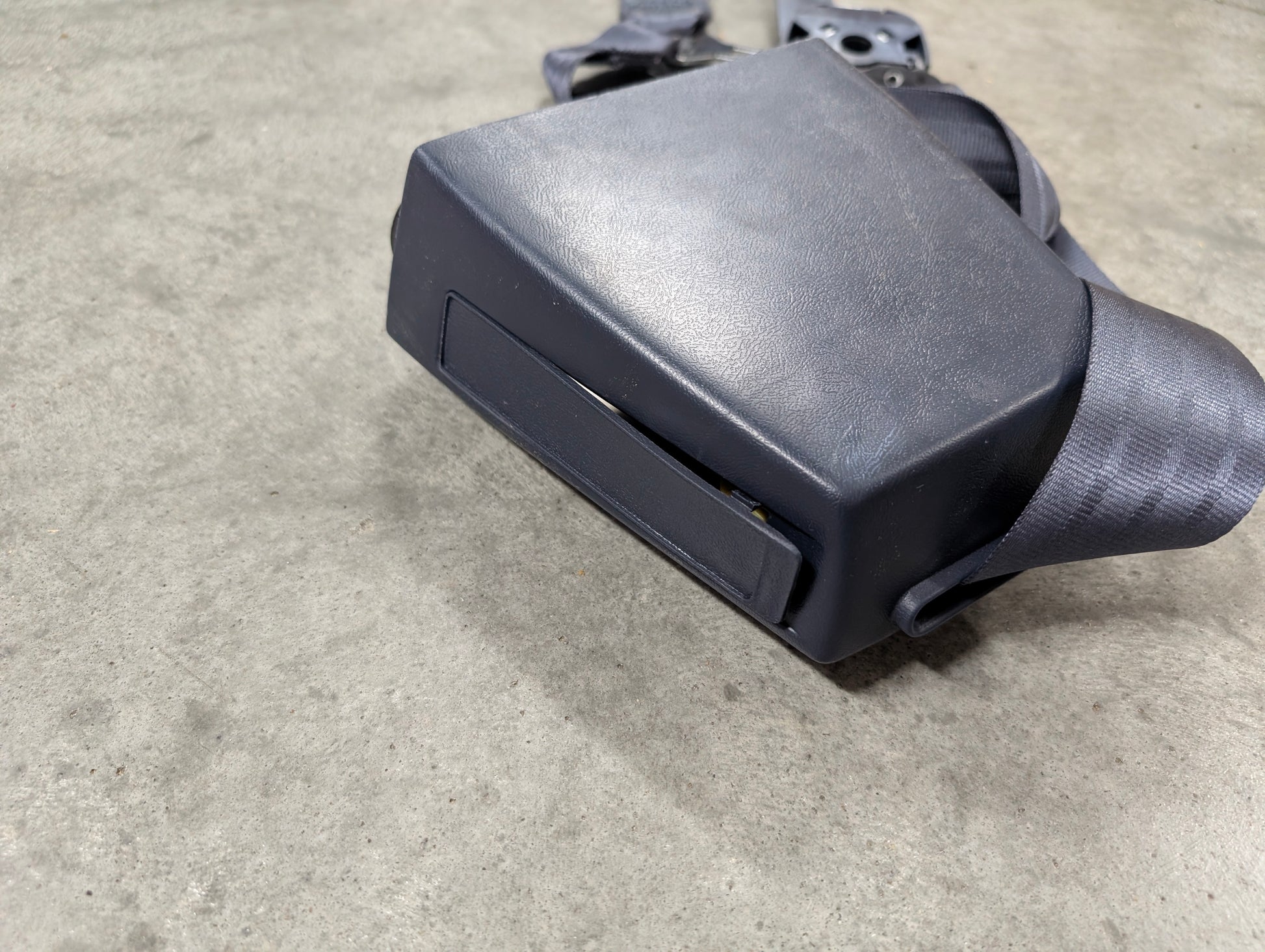 A close-up of a General Motors NOS Passenger Seat Belt Retractor & Buckle in Blue, designed for 1988-1994 Chevy/GMC C/K Trucks, resting on gray concrete. The broad rectangular buckle is ideal for seat belt replacement.