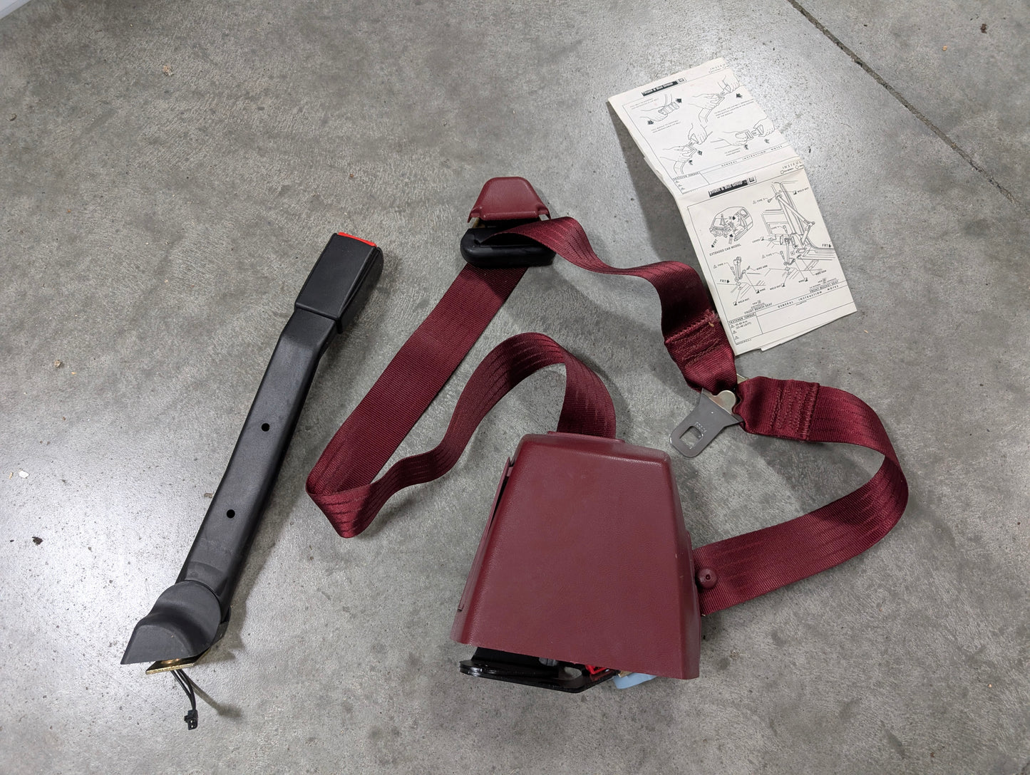 A General Motors NOS driver bucket seat belt retractor & buckle in red for 1988-1994 Chevy/GMC C/K trucks, plus an instruction manual, are arranged on a gray concrete floor.