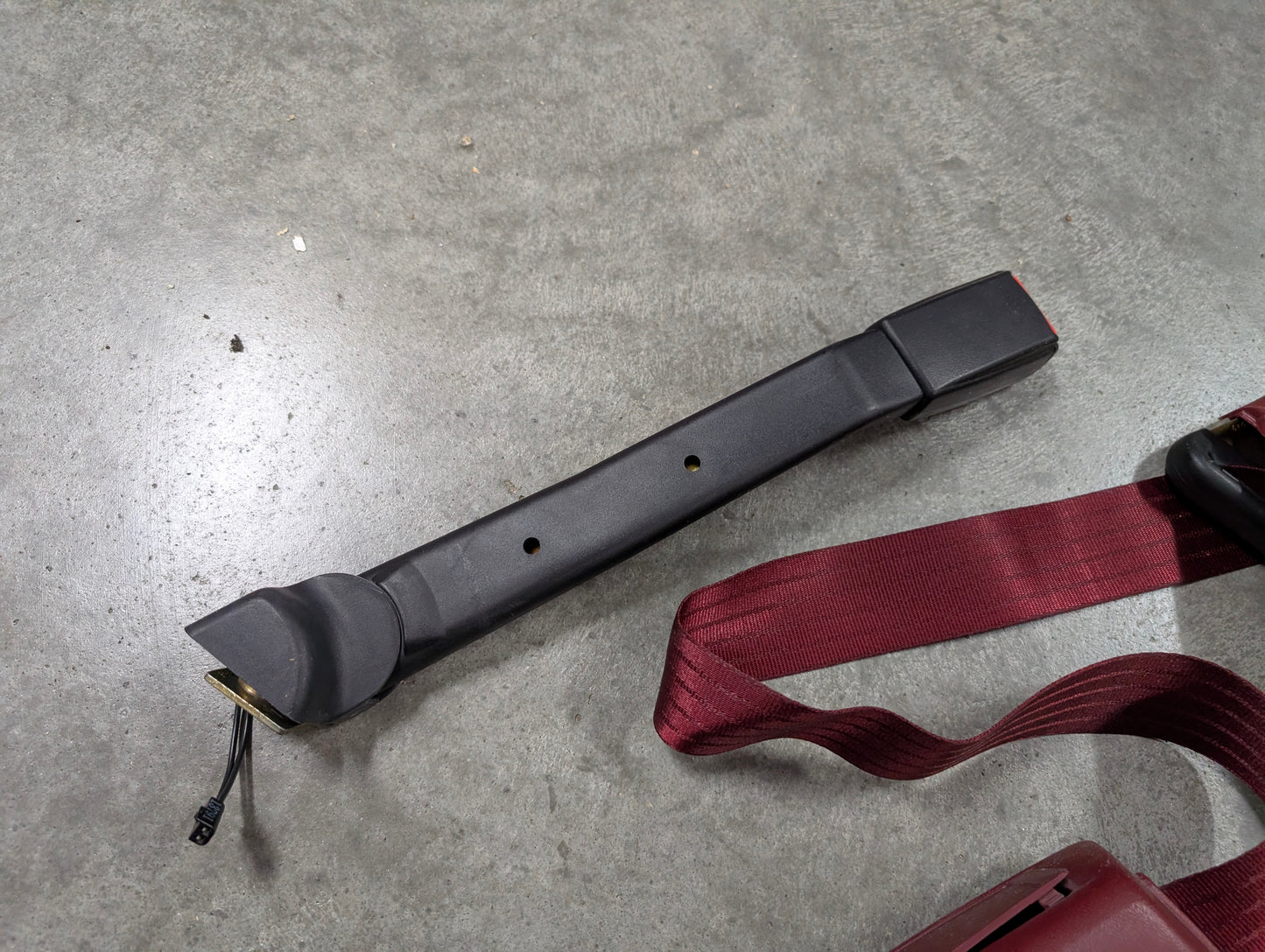 A NOS red driver bucket seat belt retractor & buckle from General Motors for 1988-1994 Chevy/GMC C/K Trucks is shown on a gray concrete surface, next to an OEM maroon seat belt.