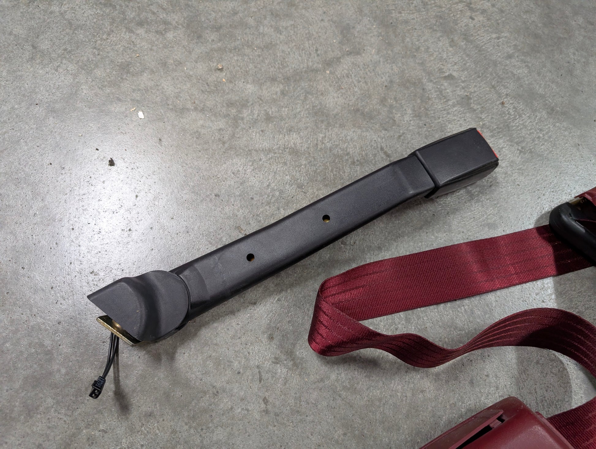 A NOS red driver bucket seat belt retractor & buckle from General Motors for 1988-1994 Chevy/GMC C/K Trucks is shown on a gray concrete surface, next to an OEM maroon seat belt.