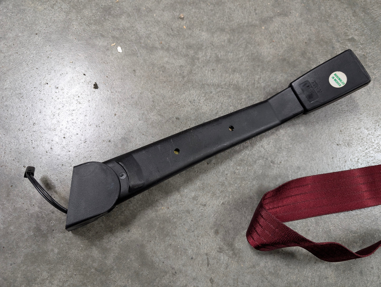 A red NOS driver bucket seat belt retractor & buckle by General Motors for 1988–1994 Chevy/GMC C/K trucks lies on gray concrete. The OEM assembly includes the retractor, buckle with short cable, and maroon seat belt strap.
