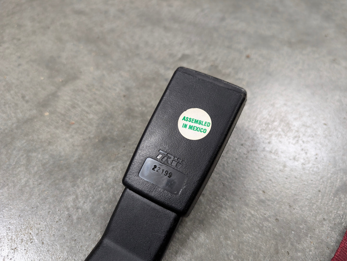 A close-up of a General Motors NOS red driver bucket seat belt retractor & buckle for 1988-1994 Chevy/GMC C/K trucks, showing an "ASSEMBLED IN MEXICO" sticker and a small label with numbers, resting on concrete.