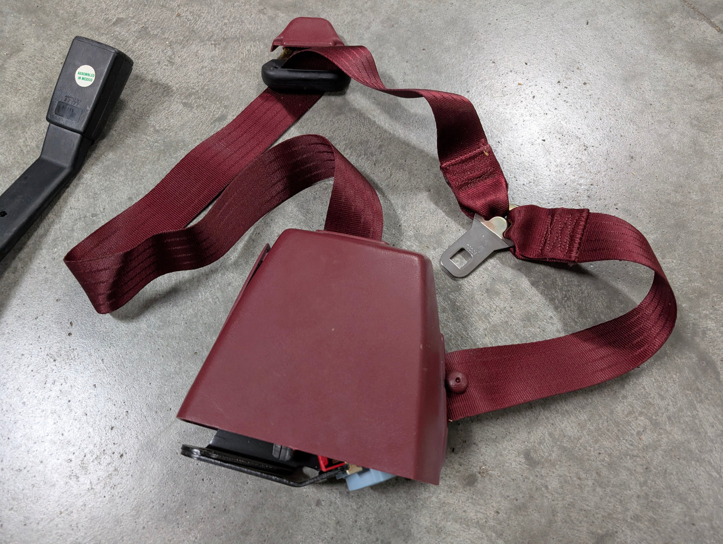 A General Motors NOS driver bucket seat belt retractor and buckle in red for 1988-1994 Chevy/GMC C/K trucks is lying uninstalled on a gray concrete surface.