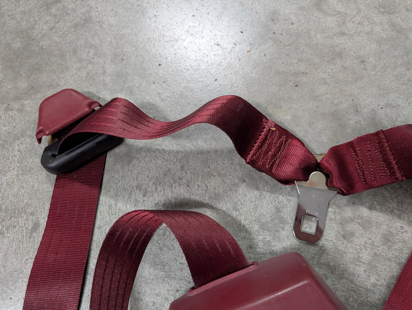 A red General Motors NOS driver bucket seat belt retractor & buckle for 1988-1994 Chevy/GMC C/K trucks is displayed on a gray textured surface, with the latch plate visible and the belt slightly twisted near the center.