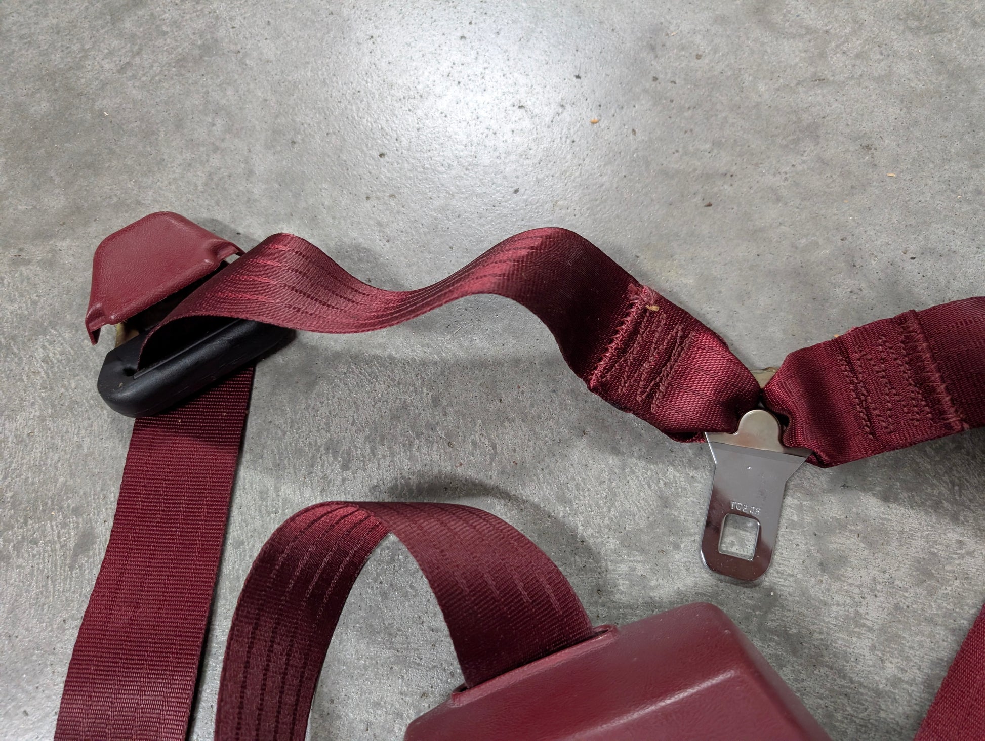 A red General Motors NOS driver bucket seat belt retractor & buckle for 1988-1994 Chevy/GMC C/K trucks is displayed on a gray textured surface, with the latch plate visible and the belt slightly twisted near the center.