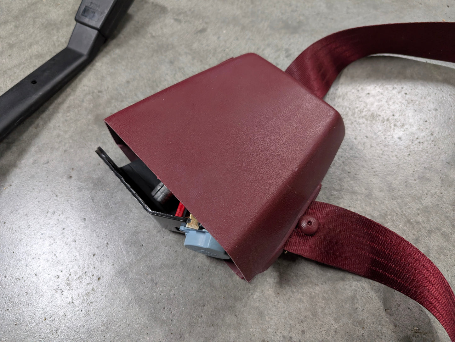 A General Motors NOS Driver Bucket Seat Belt Retractor & Buckle in red for 1988-1994 Chevy/GMC C/K Trucks rests on gray concrete, with its partially open cover displaying the seatbelt webbing and part of the metal mechanism inside.