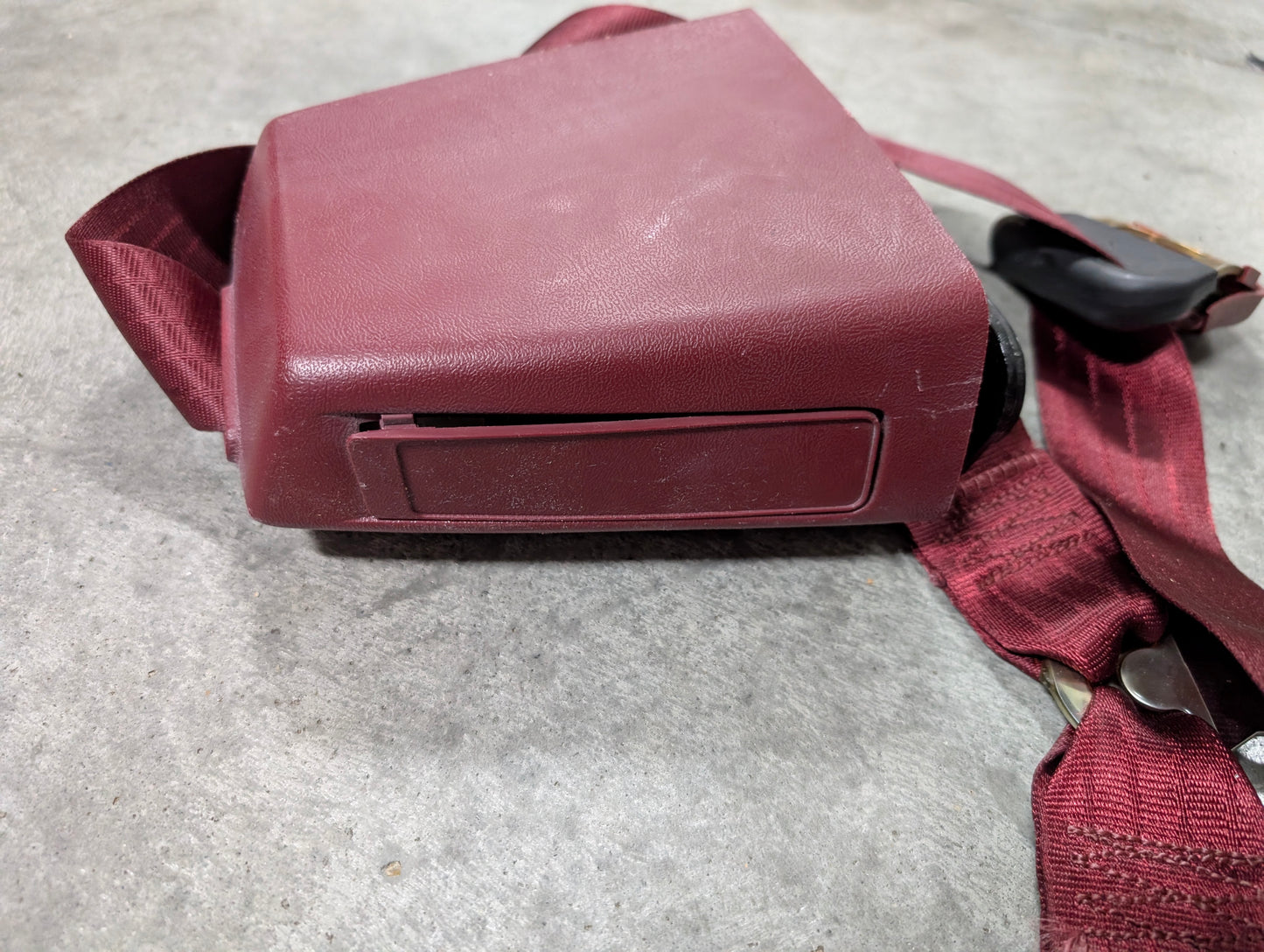 A close-up of a General Motors NOS Driver Bucket Seat Belt Retractor & Buckle in red for 1988-1994 Chevy/GMC C/K Trucks, lying on a gray concrete surface.