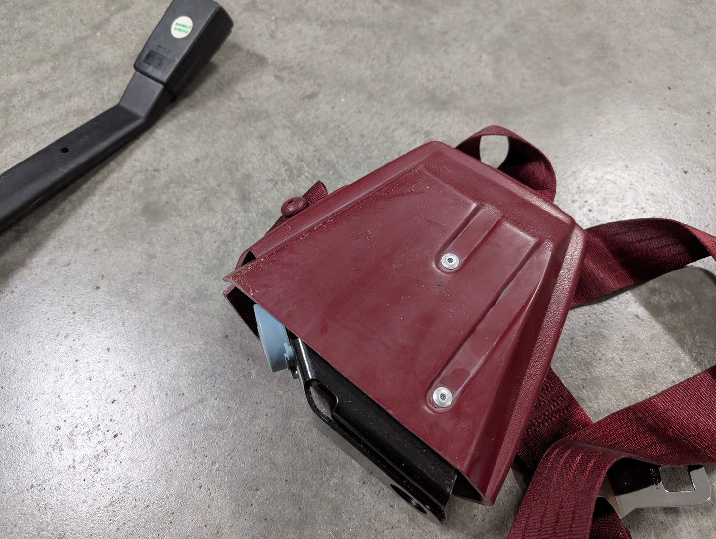 A General Motors NOS red driver bucket seat belt retractor and buckle for 1988–1994 Chevy/GMC C/K trucks rests on gray concrete, with the buckle showing a black plastic handle and green-white sticker.