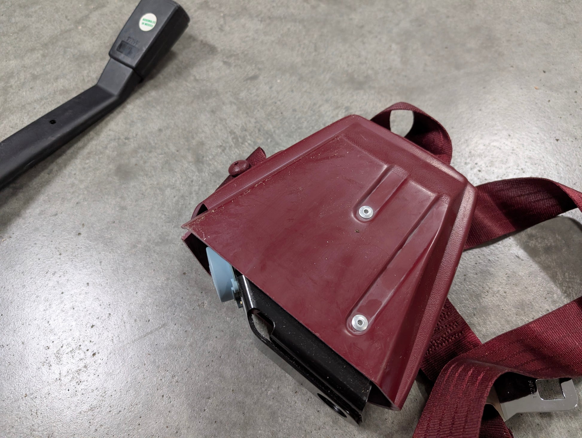 A General Motors NOS red driver bucket seat belt retractor and buckle for 1988–1994 Chevy/GMC C/K trucks rests on gray concrete, with the buckle showing a black plastic handle and green-white sticker.