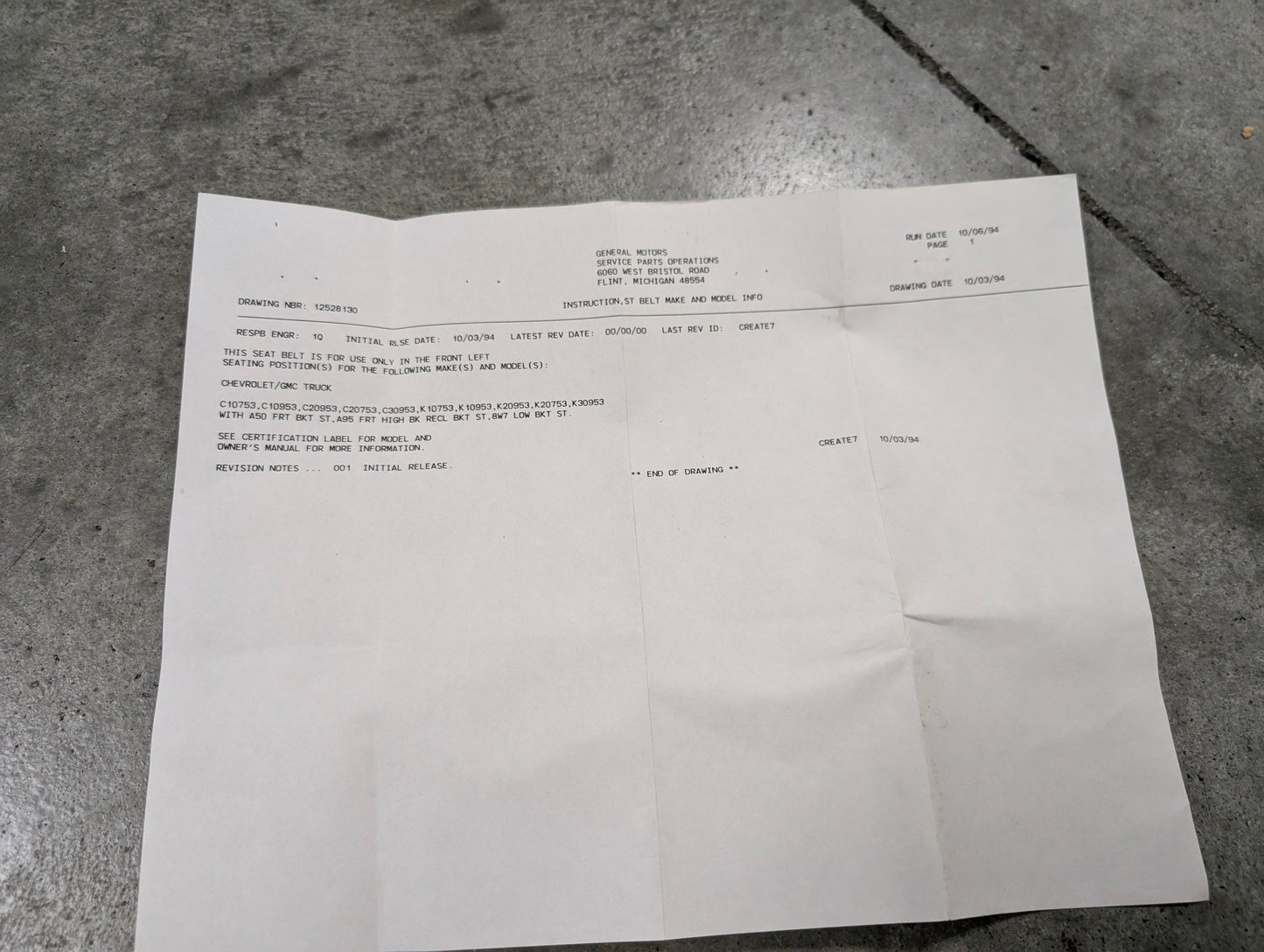 A sheet of white paper with typed text on a gray concrete floor details a General Motors NOS red driver bucket seat belt retractor & buckle for 1988-1994 Chevy/GMC C/K trucks. The document uses headings and block-style text.