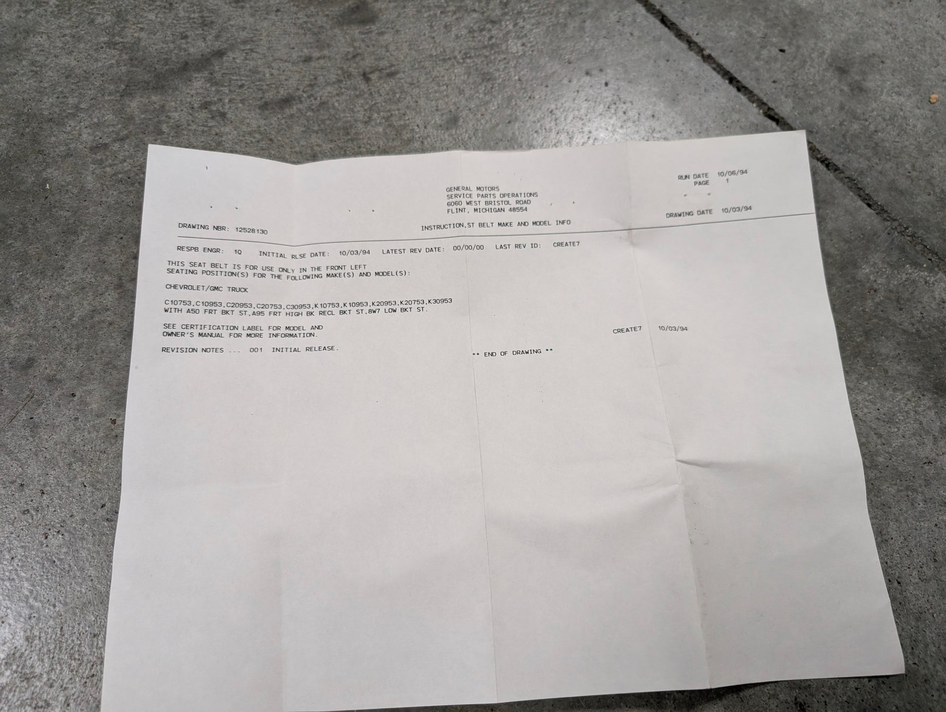 A sheet of white paper with typed text on a gray concrete floor details a General Motors NOS red driver bucket seat belt retractor & buckle for 1988-1994 Chevy/GMC C/K trucks. The document uses headings and block-style text.