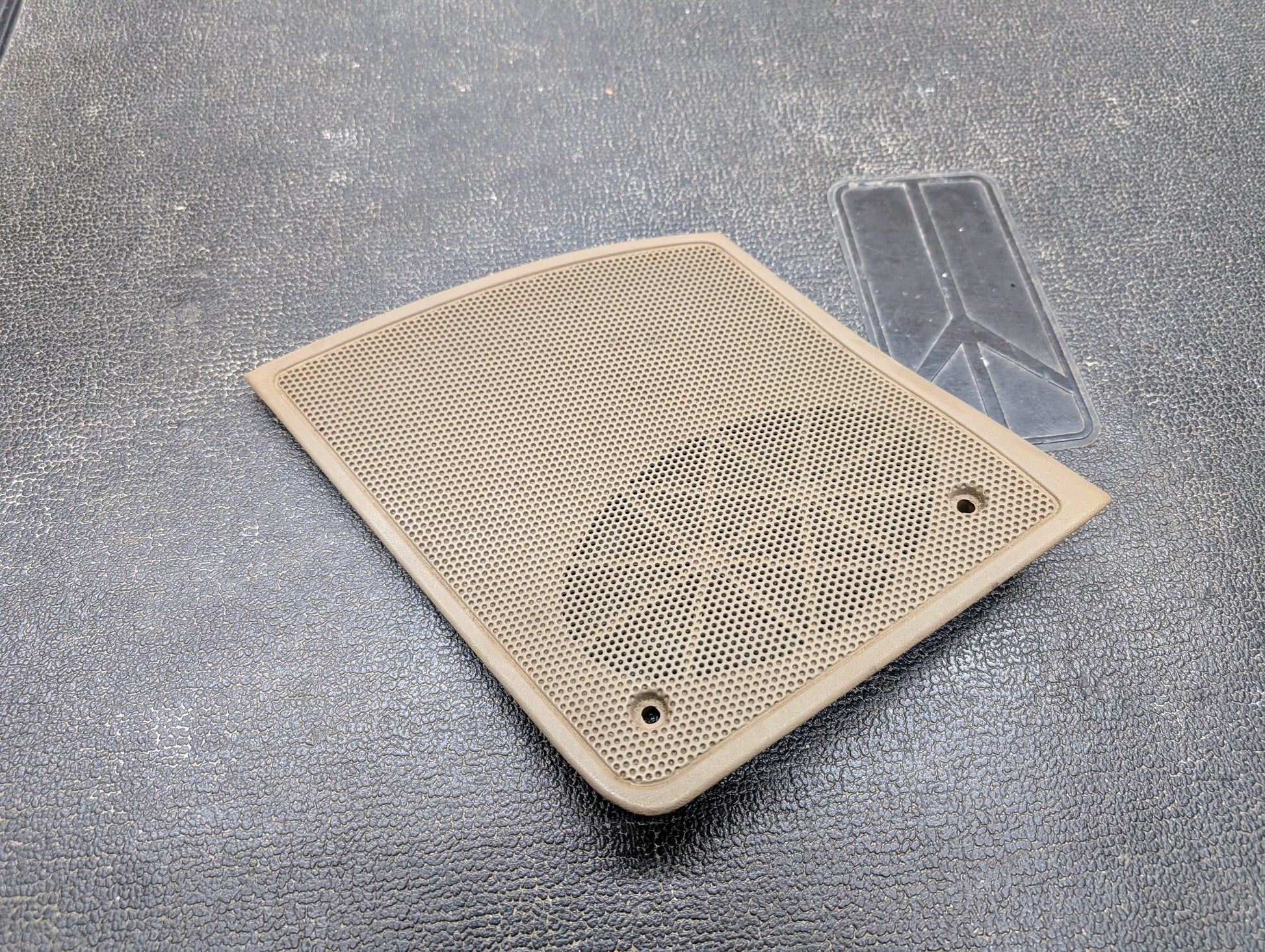 A beige OEM General Motors passenger RH dash speaker grille cover for 1995-1997 Chevrolet S10, Blazer, and more rests on a textured dark gray surface, with a faint outline of a patch nearby. Rectangular with rounded edges and two screw holes.