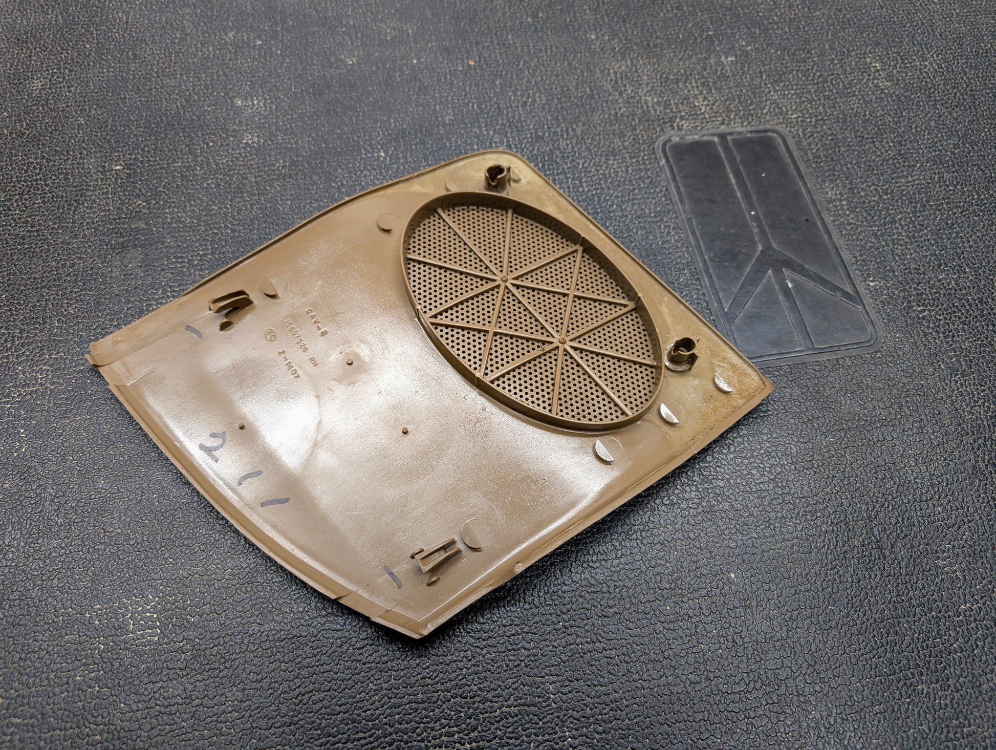 A beige/tan OEM General Motors passenger RH dash speaker grille cover for 1995-1997 Chevrolet S10, Blazer, and more rests on a textured black surface beside a clear rectangular plastic piece—perfect for interior restoration.