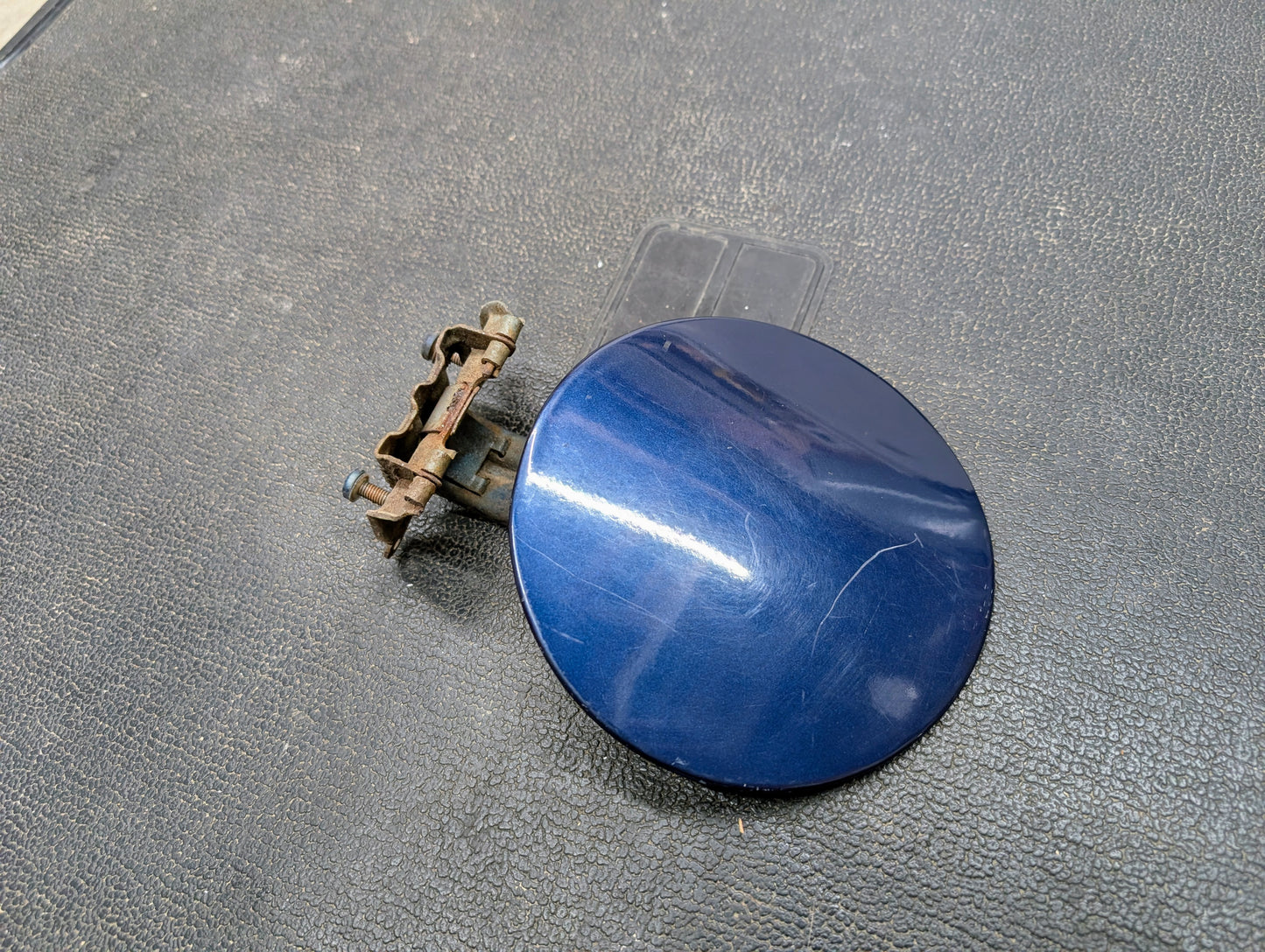 A General Motors OEM Fuel Filler Neck Door Gas Tank Cover in Blue for 2001-2004 Chevy S10 & GMC Sonoma, featuring a metal hinge and latch, sits on a textured black surface with visible scratches and a metallic reflection.
