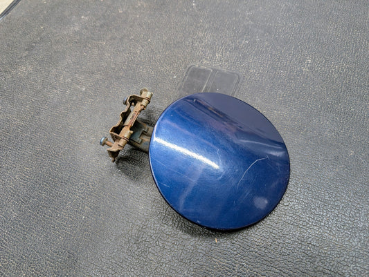 A General Motors OEM Fuel Filler Neck Door Gas Tank Cover in Blue for 2001-2004 Chevy S10 & GMC Sonoma, featuring a metal hinge and latch, sits on a textured black surface with visible scratches and a metallic reflection.