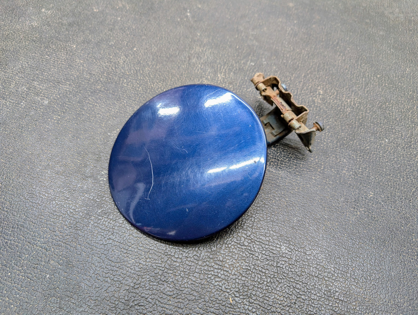 A General Motors OEM Fuel Filler Neck Door Gas Tank Cover in blue for 2001-2004 Chevy S10 & GMC Sonoma, with hinge attached, rests on a textured surface. The hinge has some rust and the cover shows a few visible scratches.