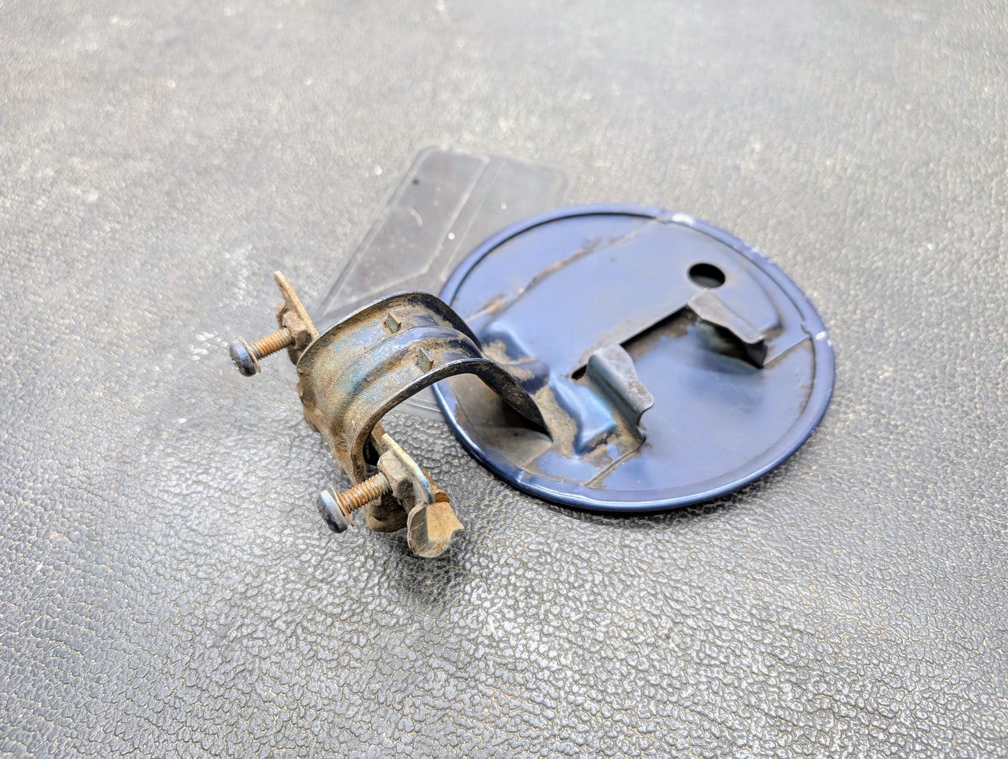 A detailed view of the General Motors OEM Fuel Filler Neck Door Gas Tank Cover in blue, made for 2001-2004 Chevy S10 & GMC Sonoma, shown with screws on a textured dark surface.