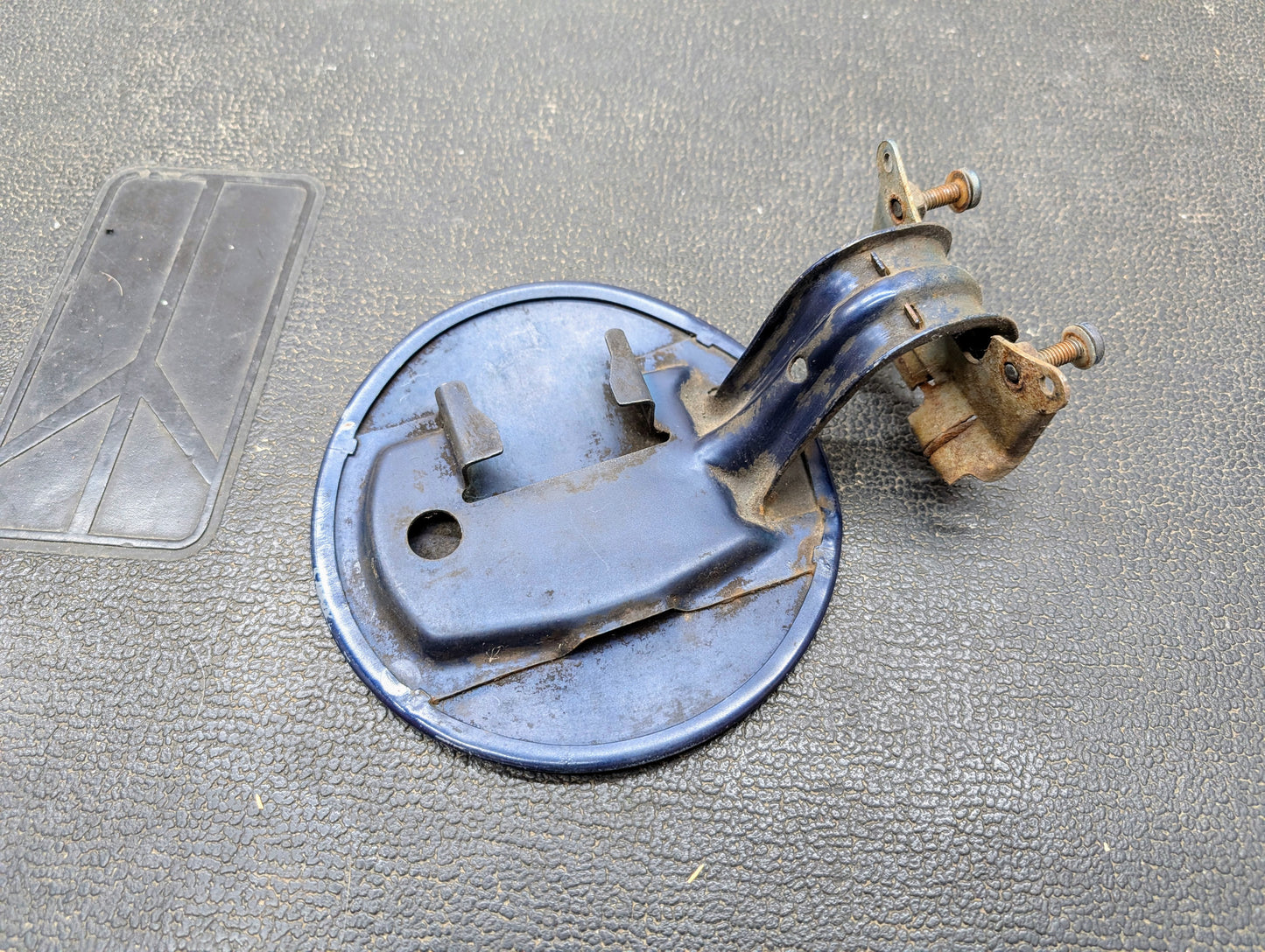 A General Motors OEM Fuel Filler Neck Door Gas Tank Cover in blue for 2001-2004 Chevy S10 and GMC Sonoma, with rusted bolts and attached mounting arm, shows wear and oxidation as it rests on a textured black surface.
