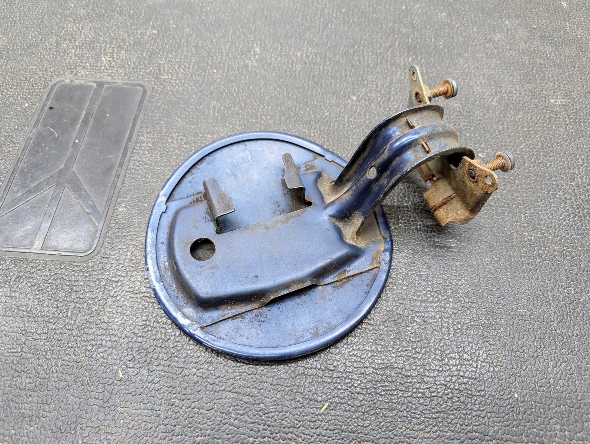 A General Motors OEM Fuel Filler Neck Door Gas Tank Cover in blue for 2001-2004 Chevy S10 and GMC Sonoma, with rusted bolts and attached mounting arm, shows wear and oxidation as it rests on a textured black surface.