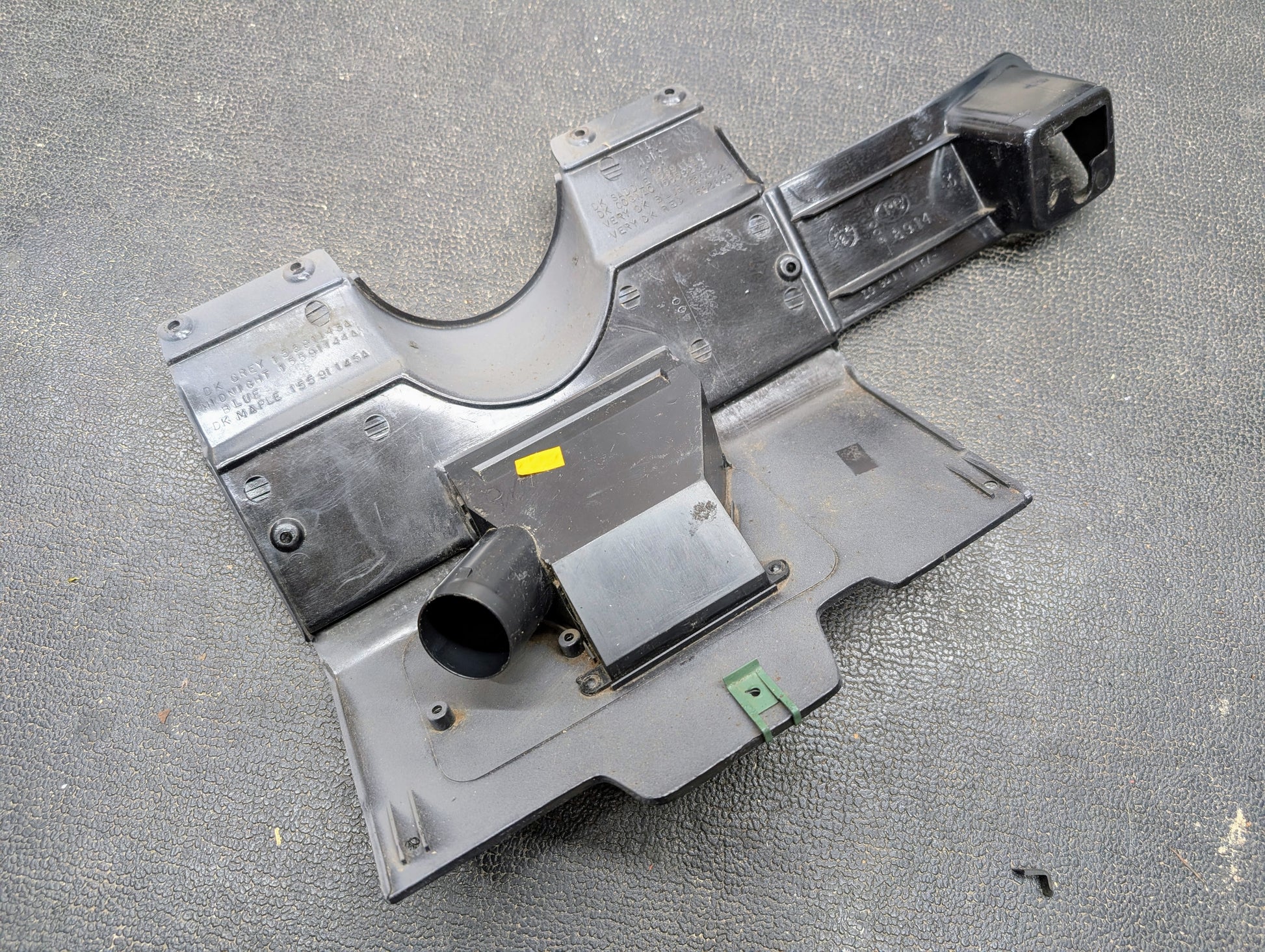 An OEM Lower Dash Trim Knee Bolster in Lt Gray for 1986-1993 Chevy S10 Blazer, GMC S15, and more by General Motors rests on a textured gray surface, featuring molded sections, mounts, a yellow sticker, and a green plastic piece.